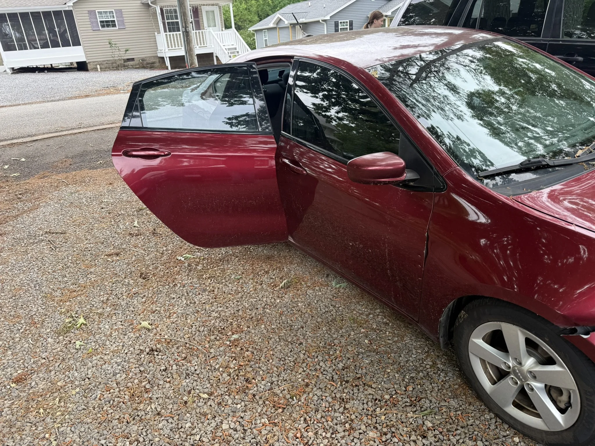 A red car parked on a gravel driveway with one of its doors open. In the background, there are houses and a woman standing near a black vehicle.