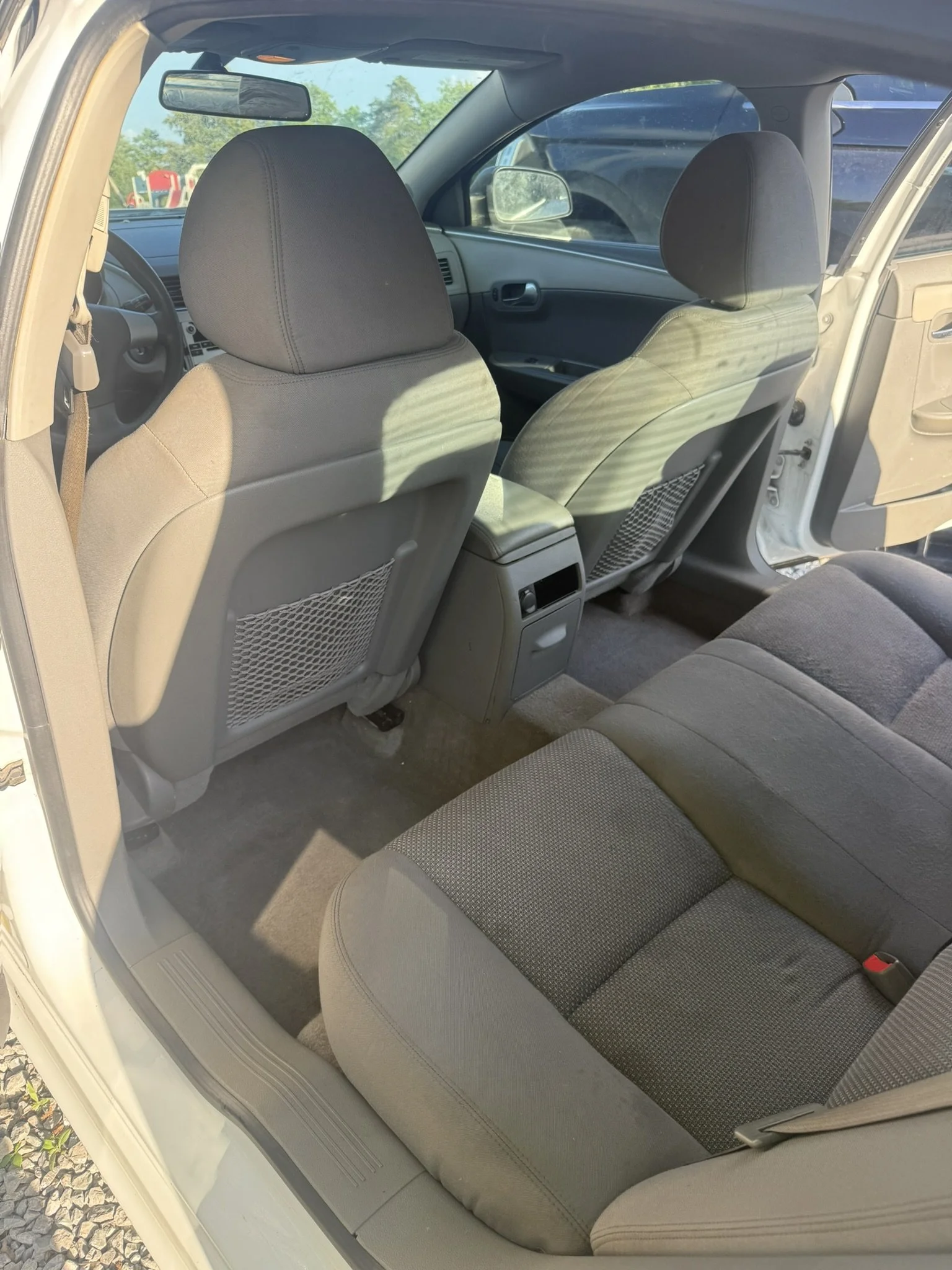 The interior of a car viewed from the open rear door, showing the back seats, front seats, and dashboard with a rearview mirror. Outside, trees and a playground are visible.