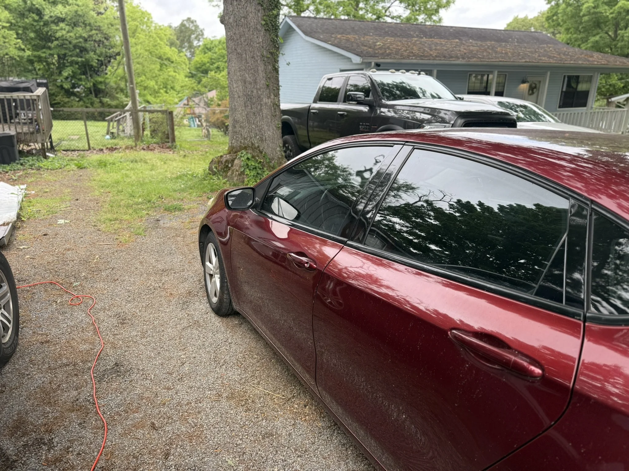 A driveway with a red car in the foreground, a black pickup truck behind it, and a house with a porch and blue siding in the background, surrounded by green trees.
