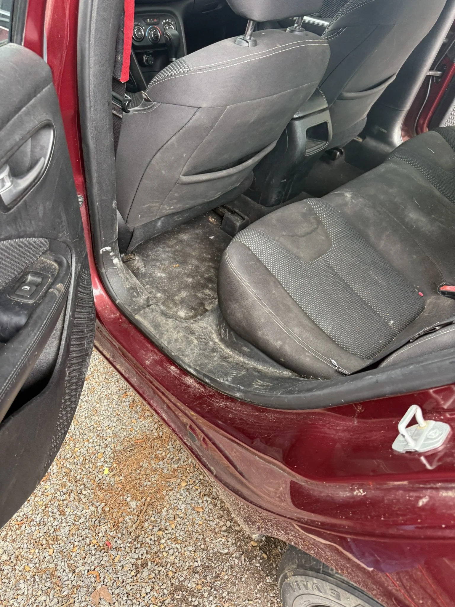 The interior of a dusty maroon car showing the back passenger side, with dirt on the floor, dirty seats, and the open door with a glimpse of the outside gravel ground.