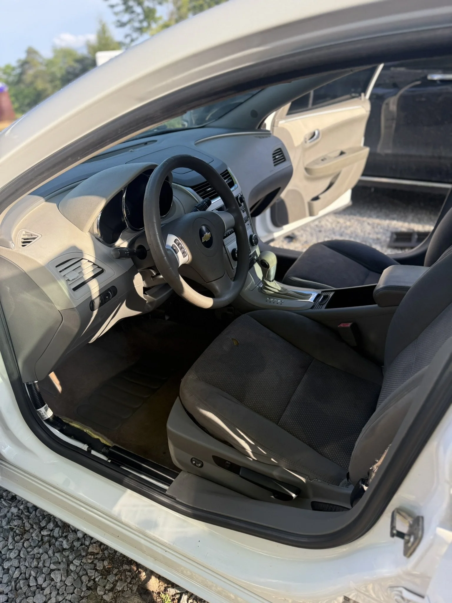 View of a car's interior from the open driver's side door, showing the steering wheel, dashboard, front seats, and part of the rear area, with a beige and gray color scheme.