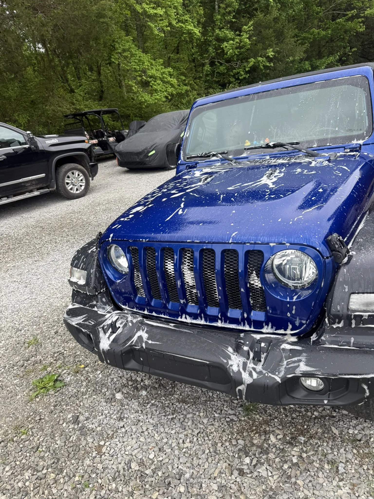 A blue Jeep with front-end damage and white foam on the hood and front bumper, parked on a gravel surface with trees in the background.