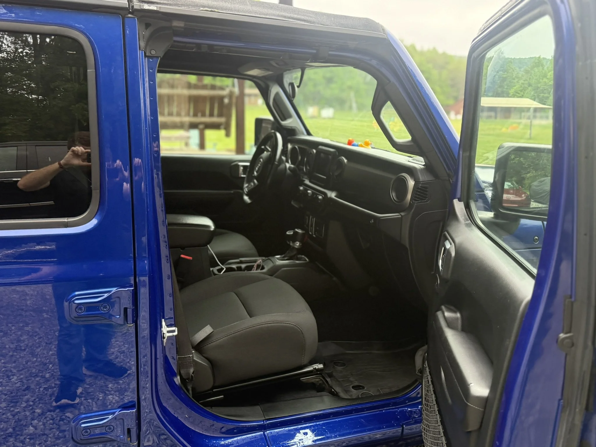 The image shows the interior of a blue vehicle, with the driver-side door open, revealing the seats and dashboard. The photo is taken outside, with a reflection of a person taking the picture visible in the window.