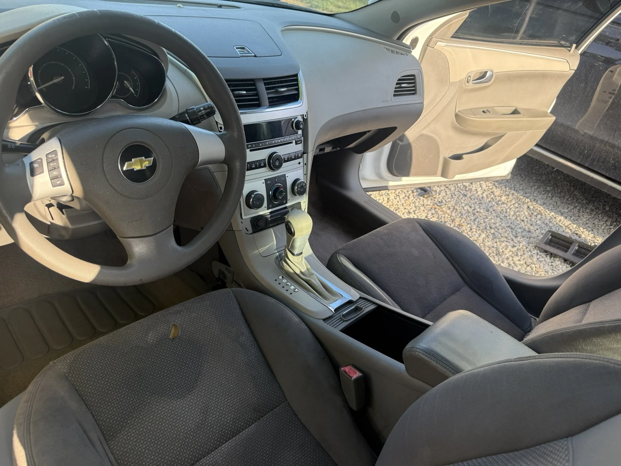 Interior of a Chevrolet vehicle, showing the steering wheel with Chevrolet emblem, dashboard, center console with controls, and front seats. The passenger side door and window are visible with sunlight coming inside.