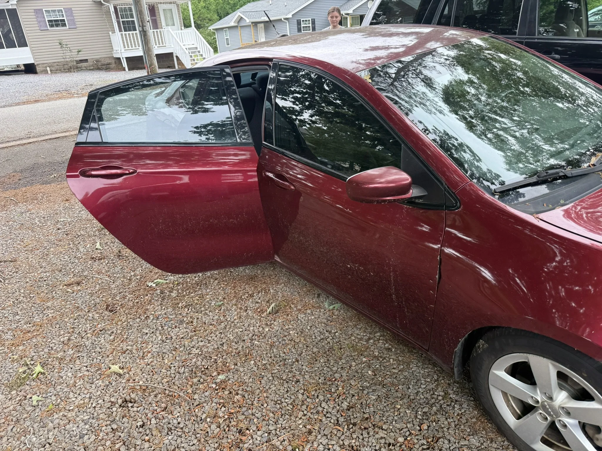 A red hatchback car with the front passenger door open, parked on a gravel driveway in a residential neighborhood. A woman with long hair is standing behind the car, with houses and trees visible in the background.