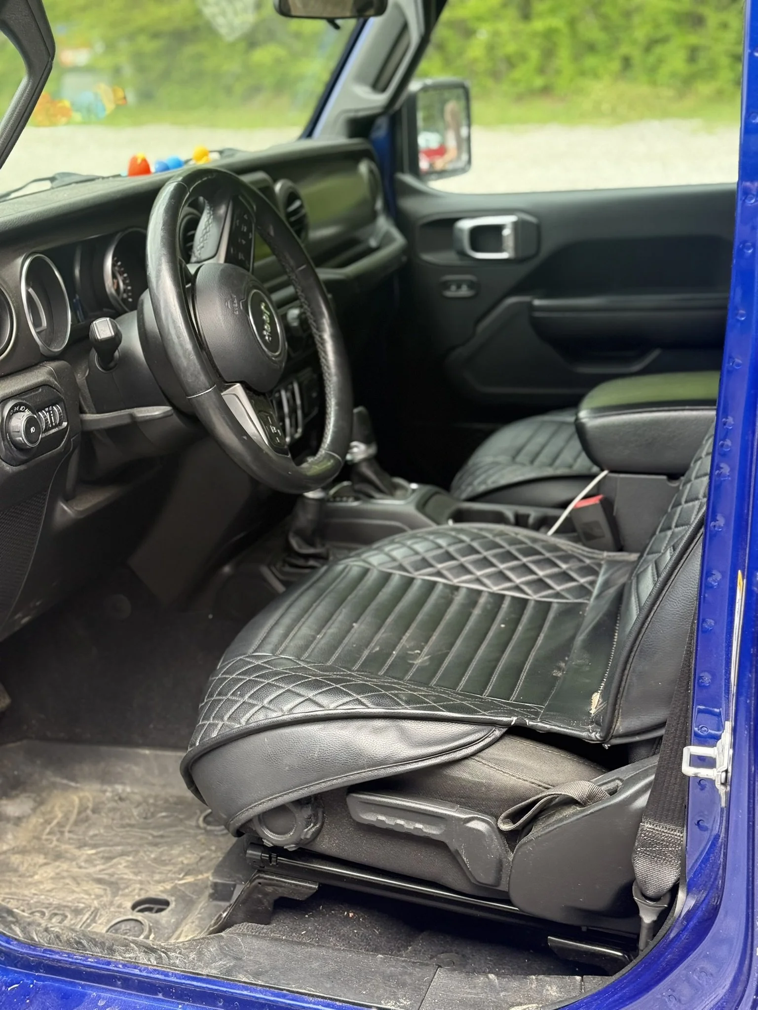 The interior of a blue Jeep vehicle showing the driver's seat with black quilted seat cover, steering wheel, dashboard, and passenger seat.