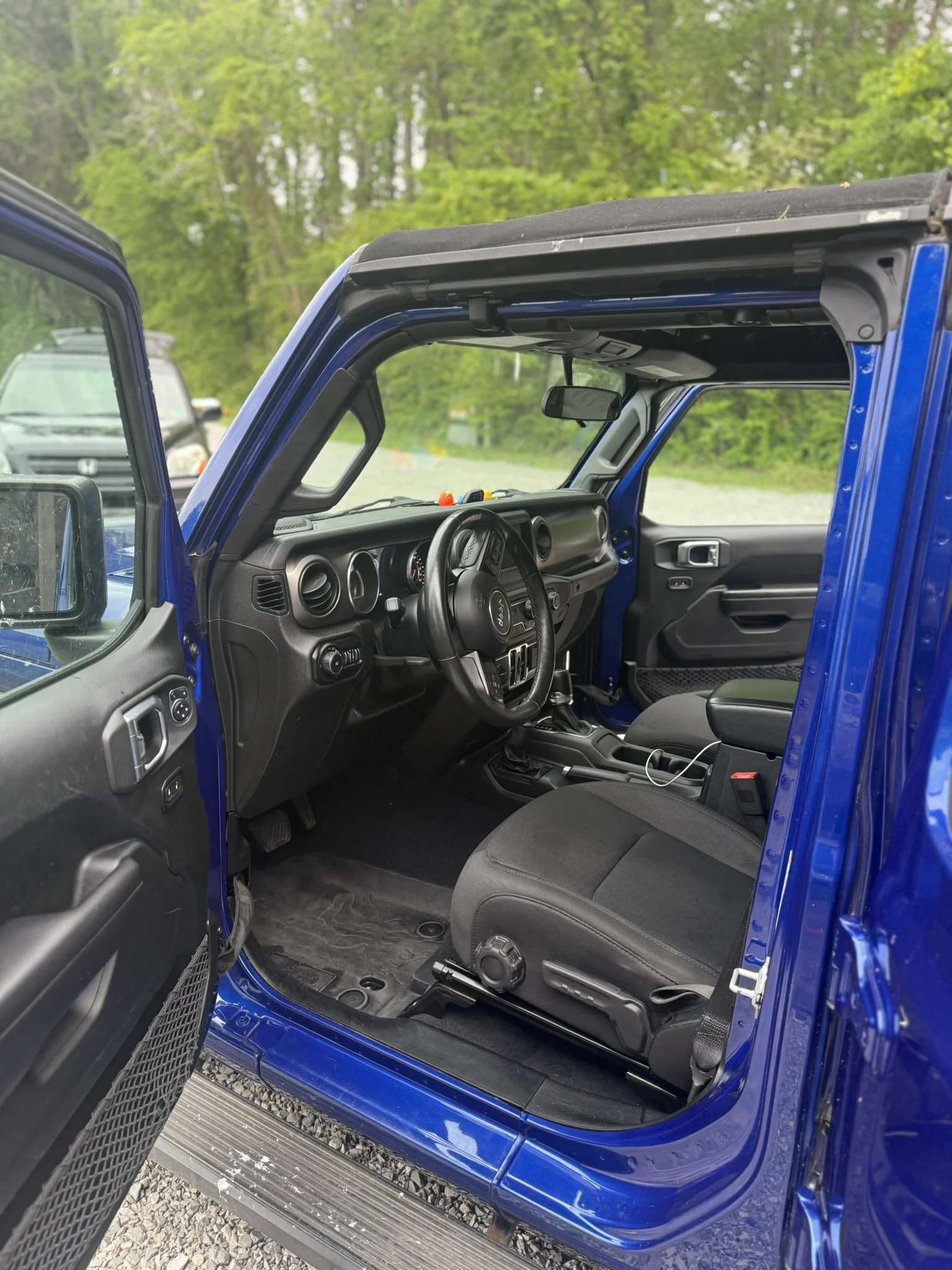 Interior of a blue Jeep with the driver's side door open, showing the dashboard, steering wheel, and front seats, with a background of trees and other cars.