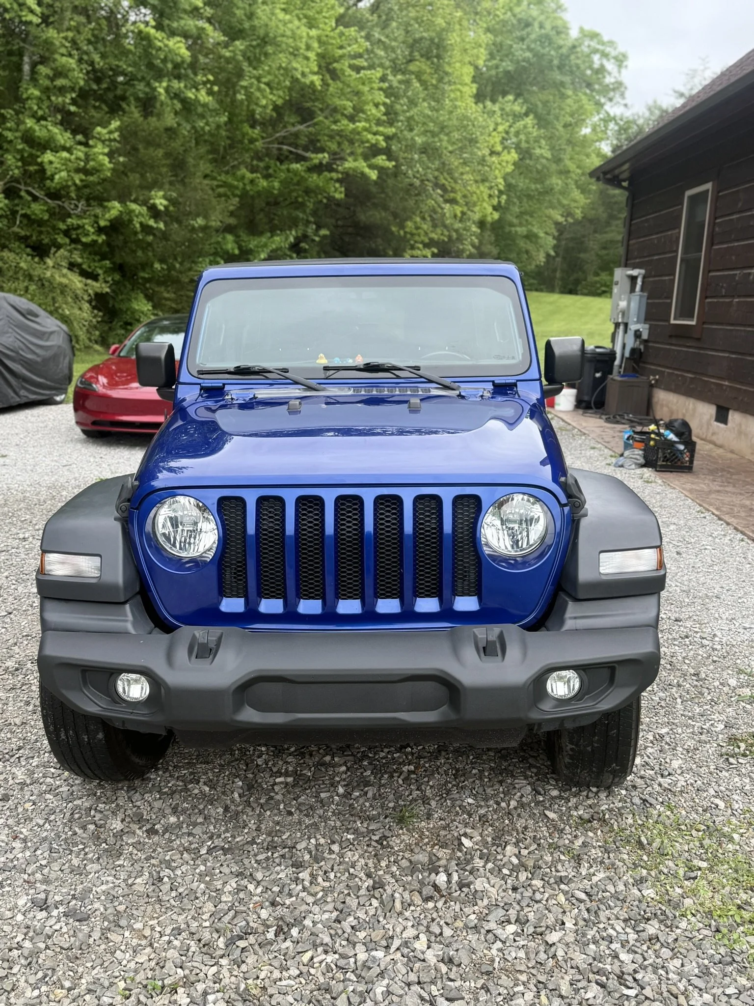 Front view of a blue Jeep vehicle parked on a gravel driveway, with a red sports car and a covered object to the left, and a wooden house and green trees in the background.