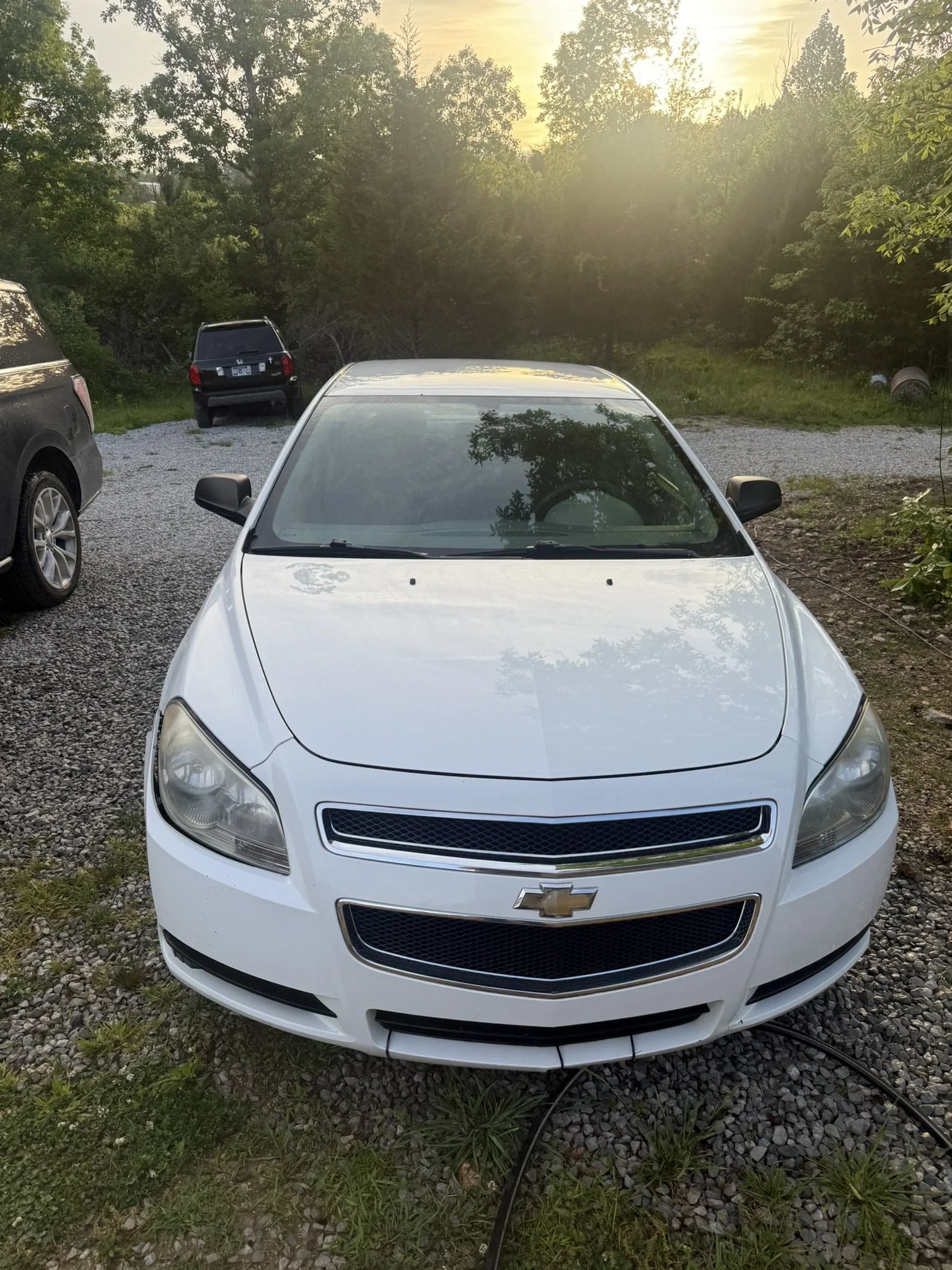 White Chevrolet car parked on a gravel driveway with trees and a sunset in the background.