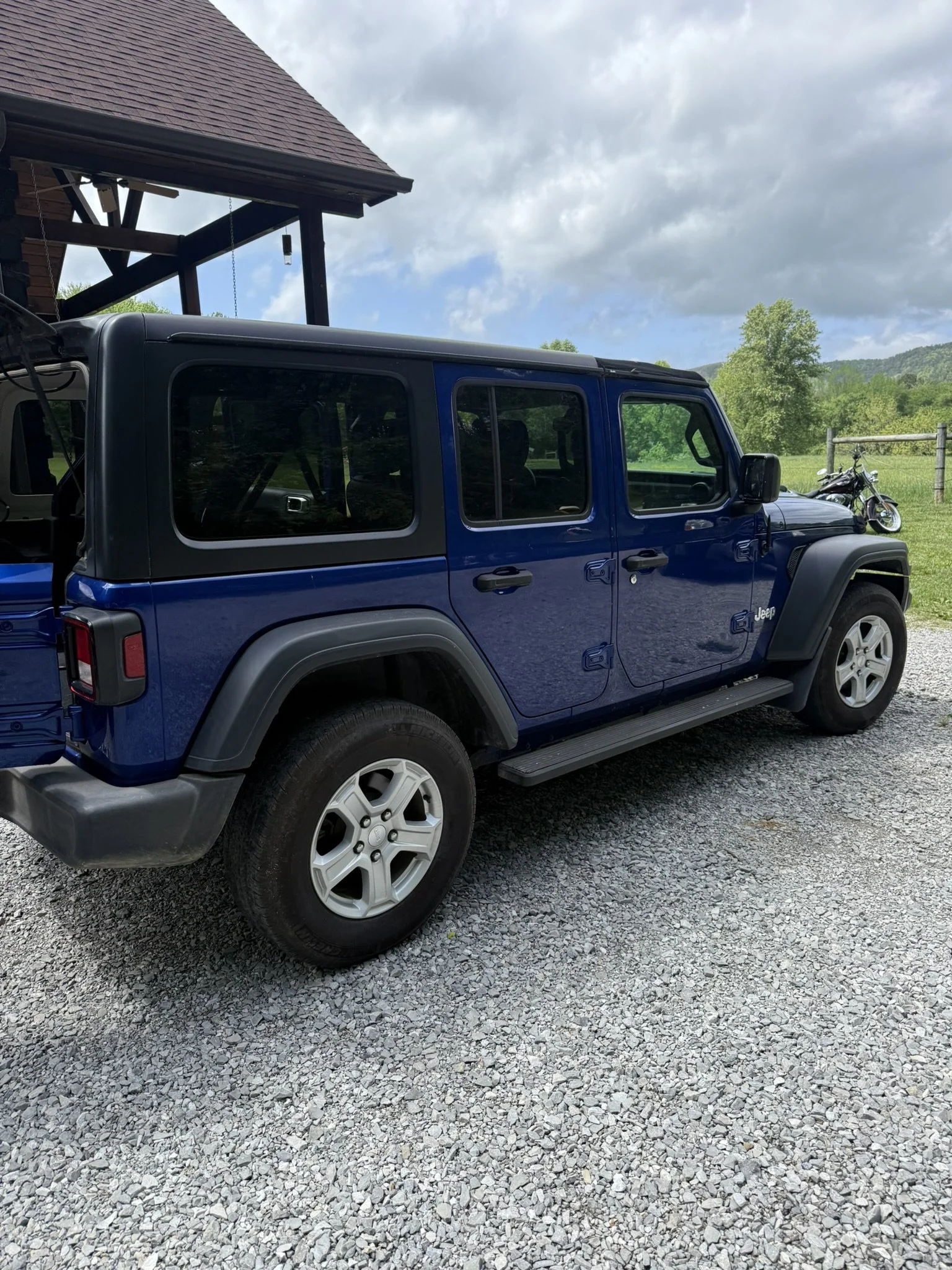 A blue Jeep parked on a gravel driveway near a wooden house with a porch and swing, with a motorcycle in the background and green trees under a partly cloudy sky.