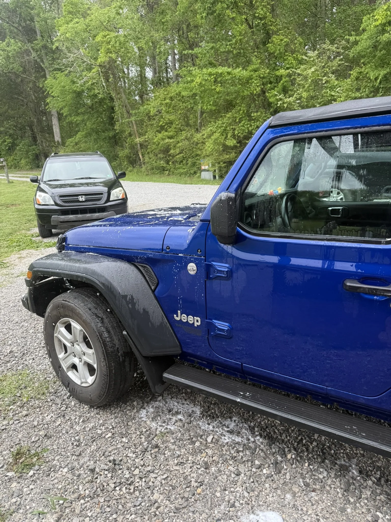 A blue Jeep and a black Honda SUV parked on a gravel driveway next to a wooded area with green trees and grass.