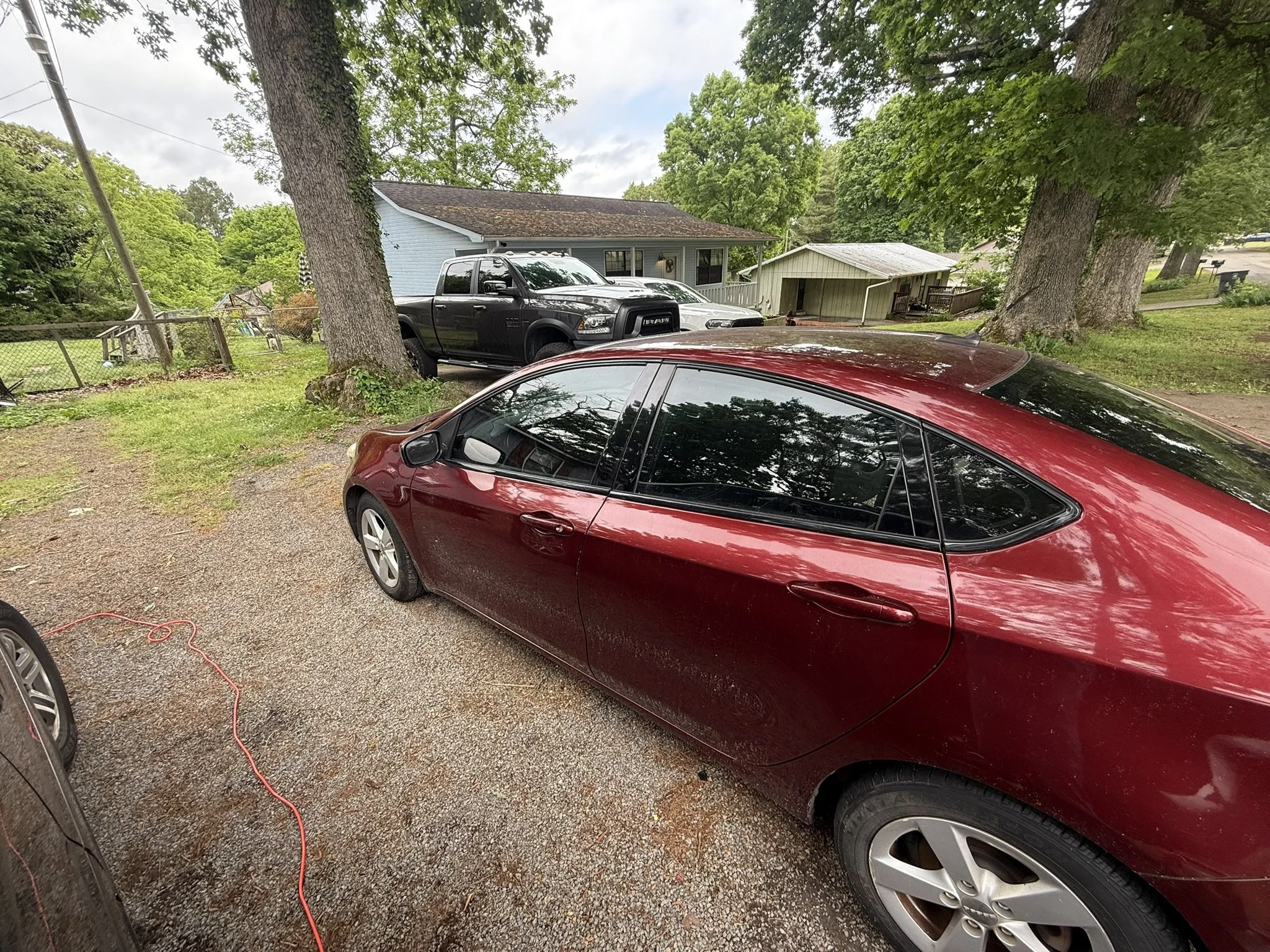 Residential driveway with red, black, and gray vehicles parked under large trees, in front of houses with green lawns.