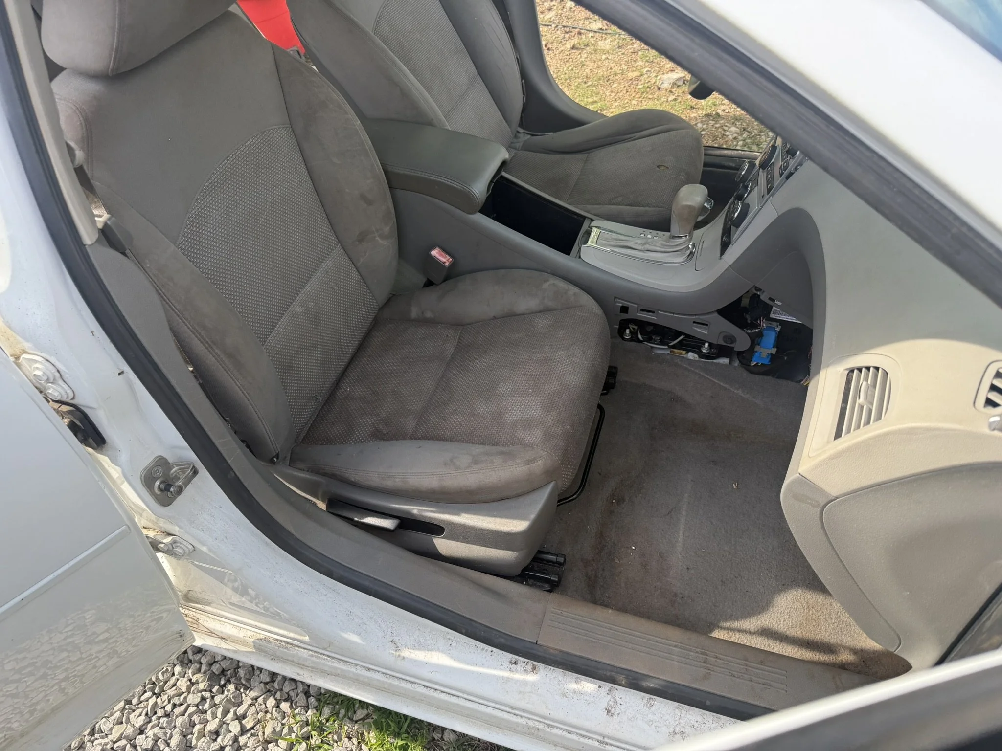 The image shows the interior of a grey car with the driver's side door open, revealing two front seats, the center console, and the gear shift. The dashboard and various controls are partially visible.