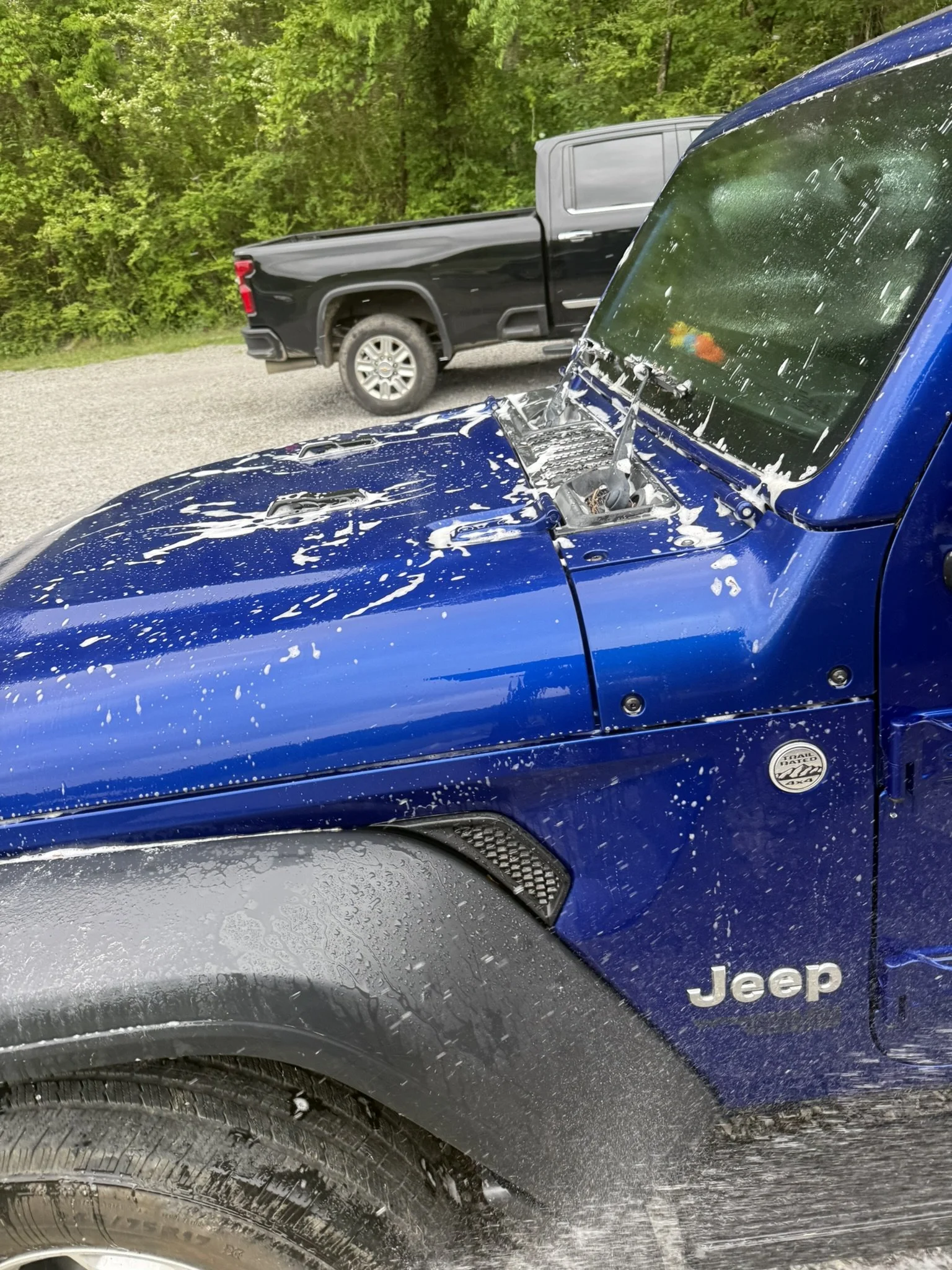 A close-up of a blue Jeep truck with white splashes of mud and water on the hood and side, parked outdoors with a second black pickup truck and dense green foliage in the background.