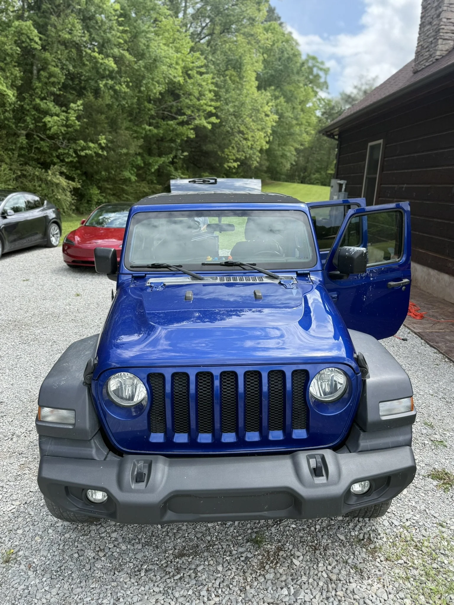 Front view of a blue Jeep in a gravel driveway, with a black house to the right and multiple parked cars to the left, surrounded by green trees and blue sky.