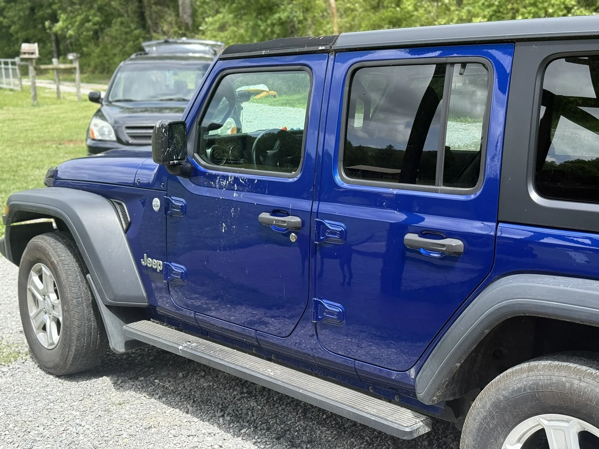 Close-up of a blue Jeep Wrangler with dirt and scratches on the side, parked on gravel with other vehicles and greenery in the background.