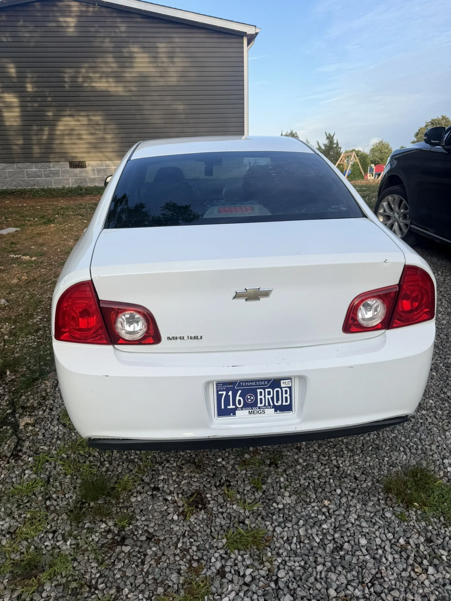 White Chevrolet Malibu car parked on gravel with a Tennessee license plate, next to a black vehicle, during daytime with a partly cloudy sky and a playground in the background.