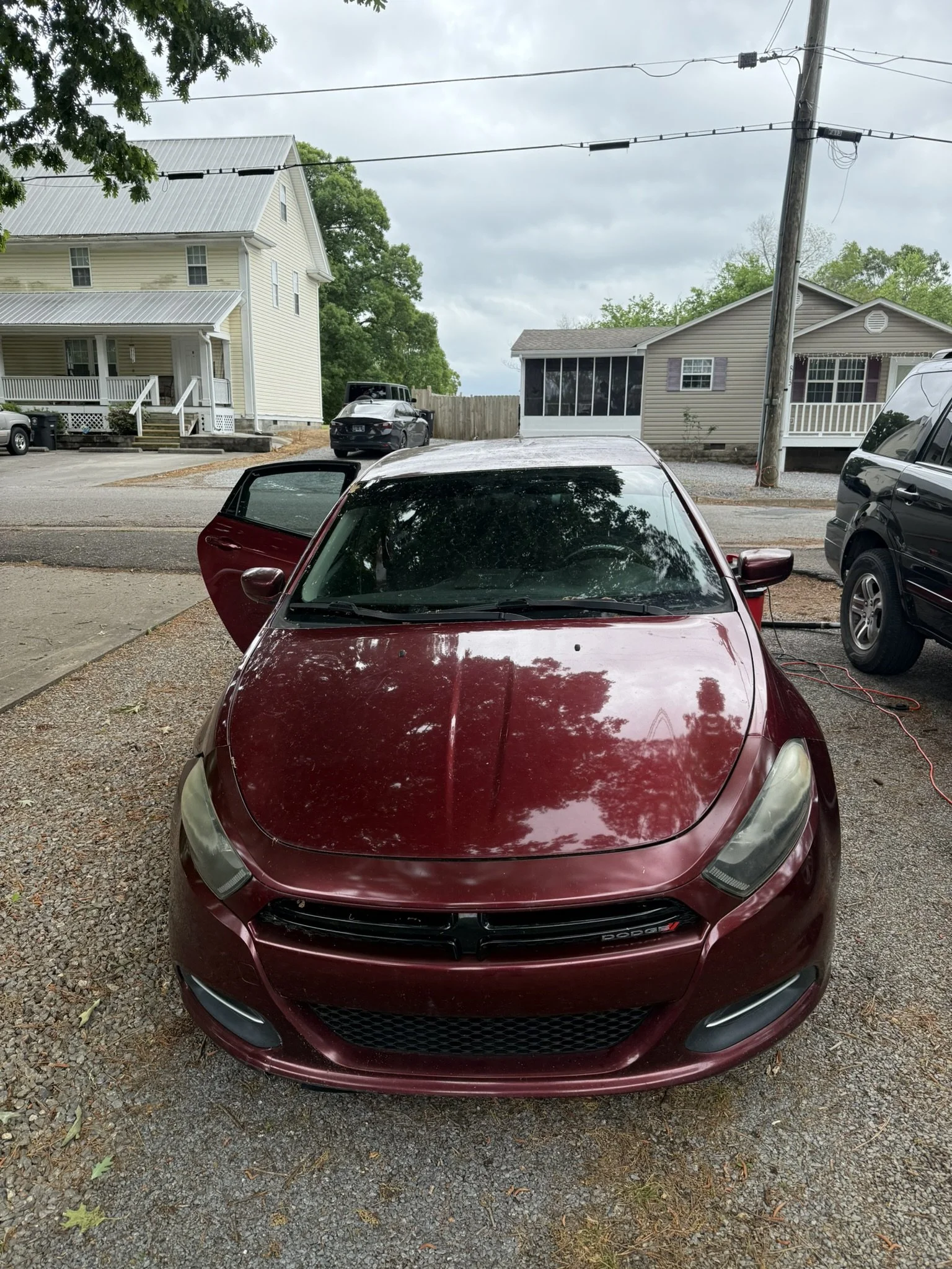 Red car with open door parked on gravel driveway in front of houses with trees and power lines in the background.