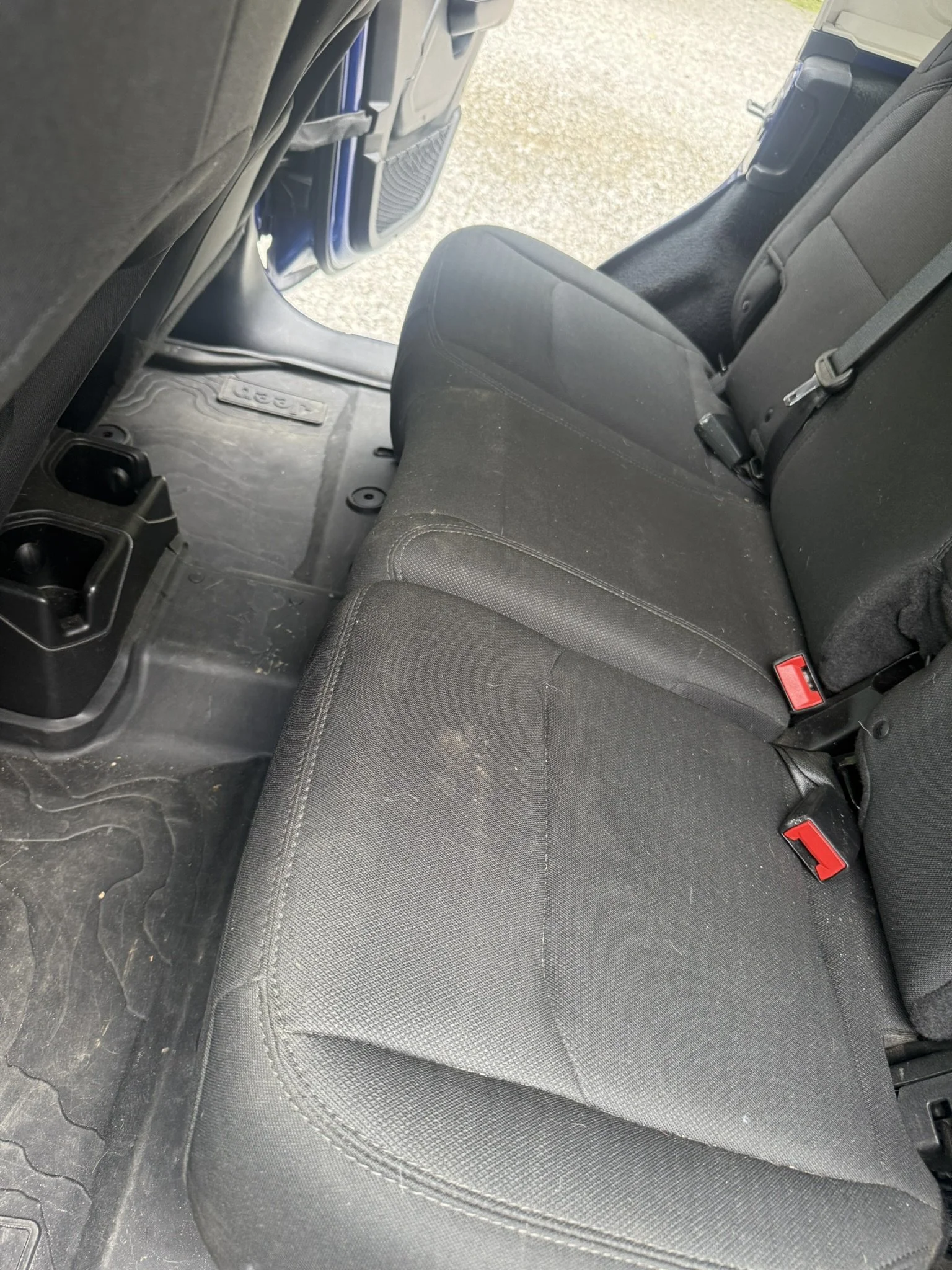Inside view of a vehicle's backseat area, showing black fabric seats, rear seat belts, a floor mat, and the back of the front seat with a seatback pocket.