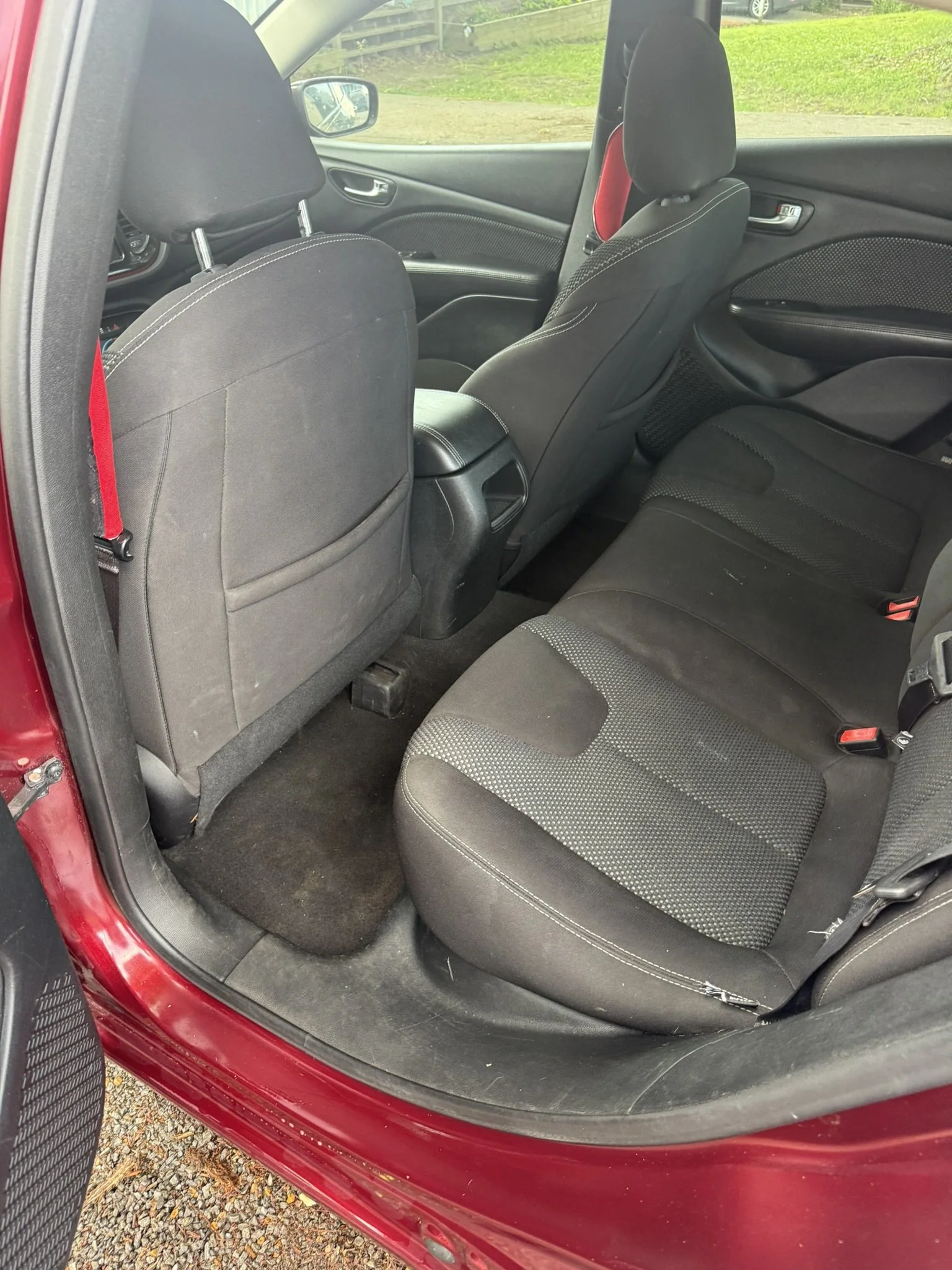 Backseat area of a red car showing black fabric seats, headrests, door panel with handle, and part of a gravel ground outside.