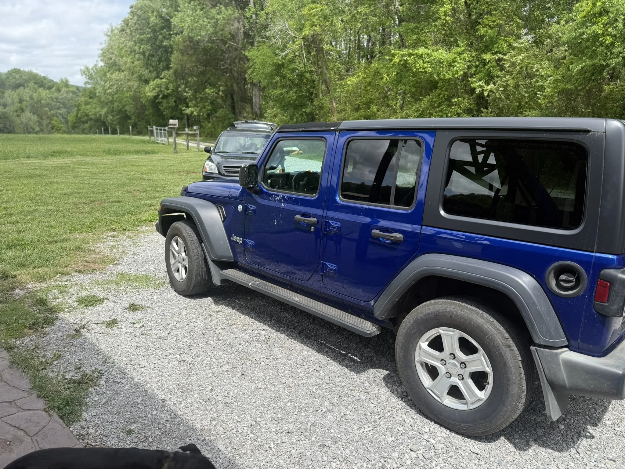 A blue Jeep SUV parked on a gravel area next to a grassy field with a black vehicle in the background. Tall trees with green leaves are along the edge of the field, under a cloudy sky.