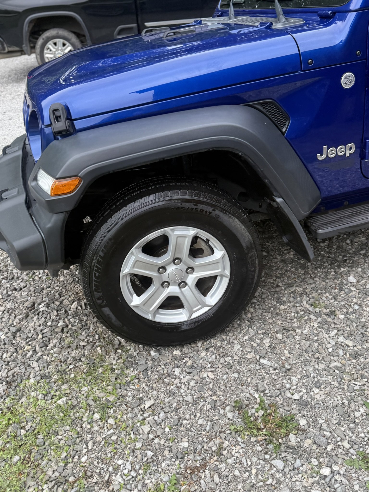 Close-up of a blue Jeep Wrangler front left wheel and fender, with a gravel ground and another vehicle in the background.
