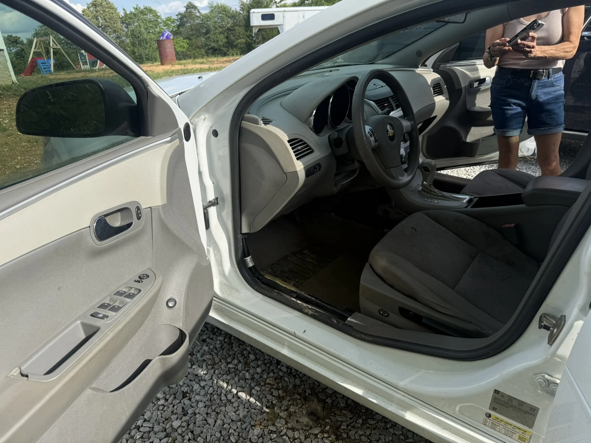 View of a white car's interior with the door open, showing the steering wheel, dashboard, and front seats. A woman in a sleeveless shirt and denim shorts is standing outside, using her phone. The background includes outdoor greenery, trees, and playg