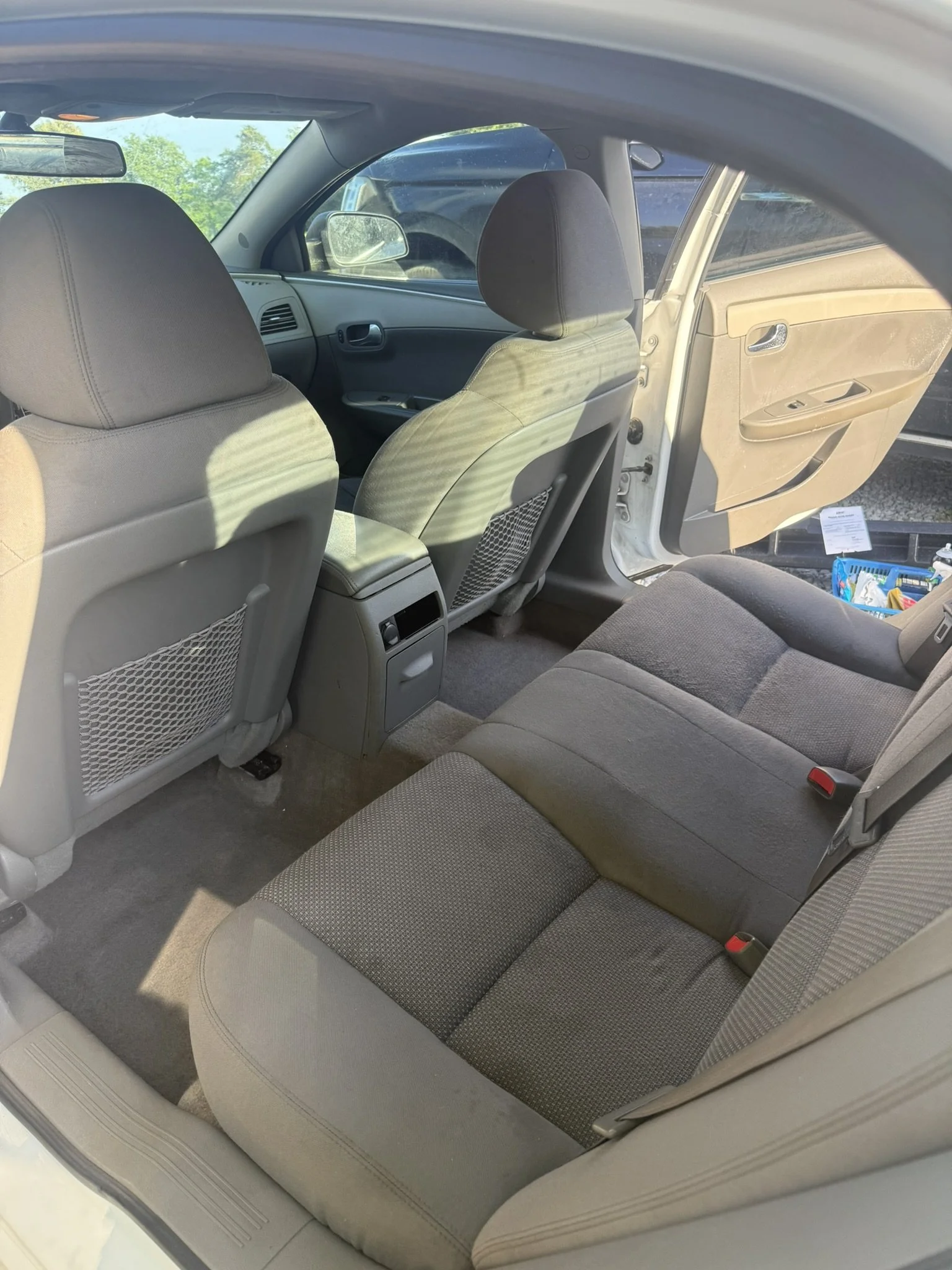 The interior of a car showing the back seat and front seats, with the open door revealing the outside. The seats are covered with gray fabric, and there is a blue basket with items on the floor near the door.