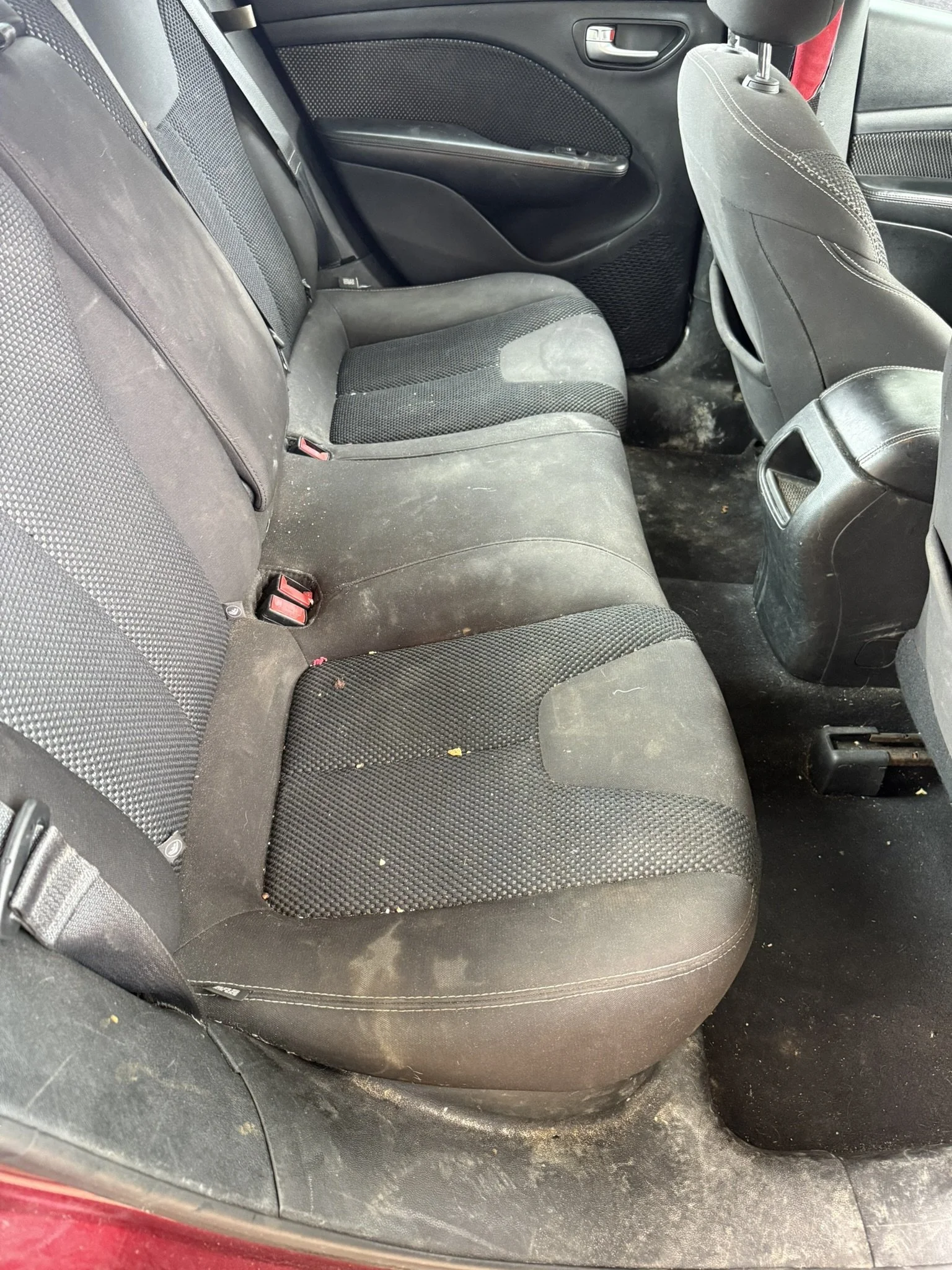 The back seat of a car with black fabric upholstery, showing some dirt and debris on the seat and floor.