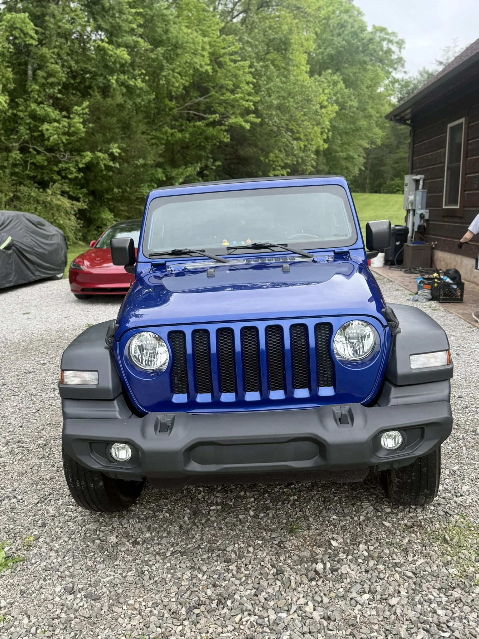 A blue Jeep parked on a gravel driveway in front of a house with wooden exterior, with a red sports car and a covered vehicle nearby, and green trees in the background.