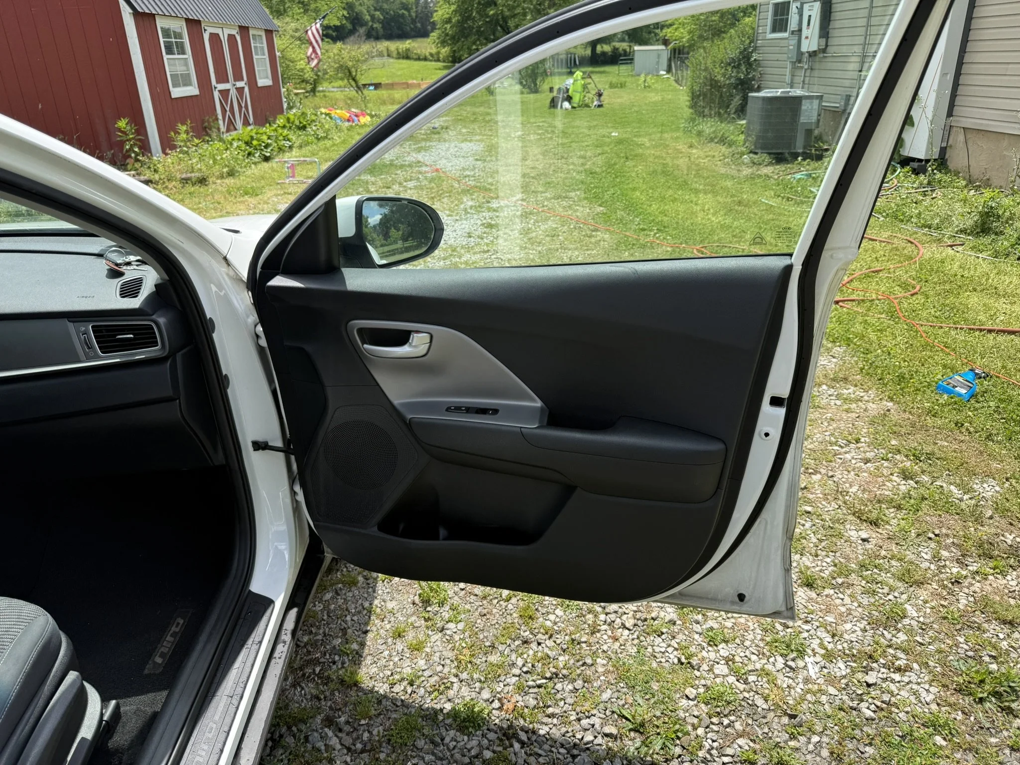 View from inside a car with the passenger side door open, showing a grassy backyard with a red shed, a flagpole with an American flag, a toy or small garden tractor, a house with electrical equipment outside, and garden tools. The ground outside has 