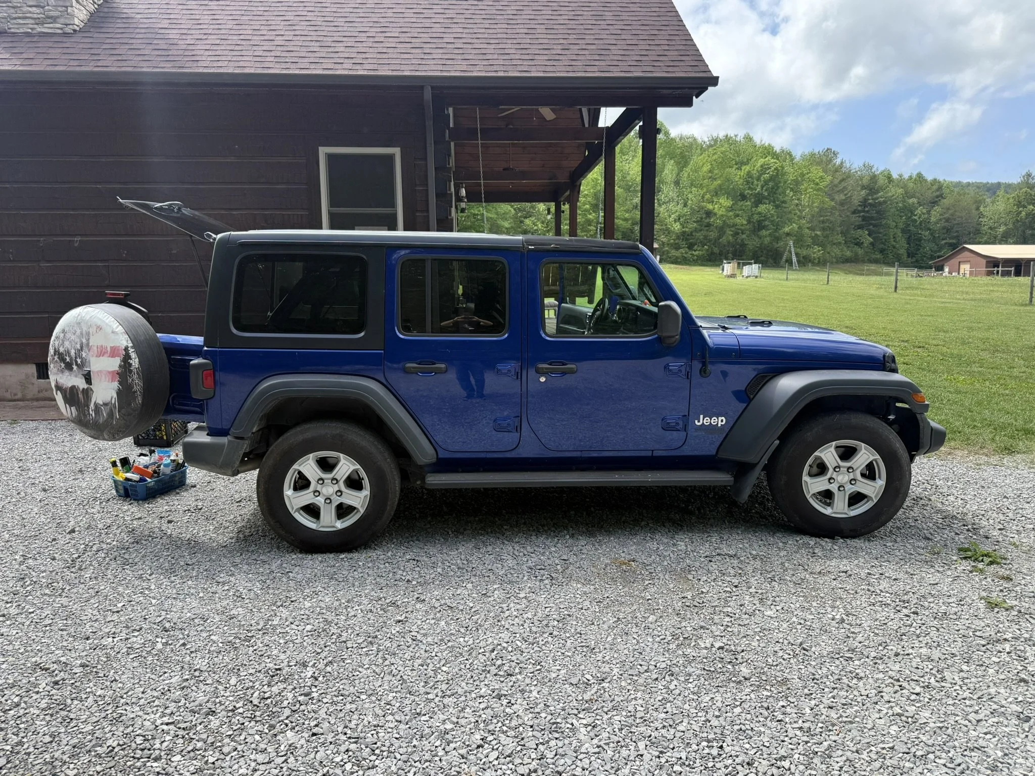 Blue Jeep Wrangler parked on gravel driveway next to a brown house with a screened porch, in a rural area with green trees and a cloudy sky.