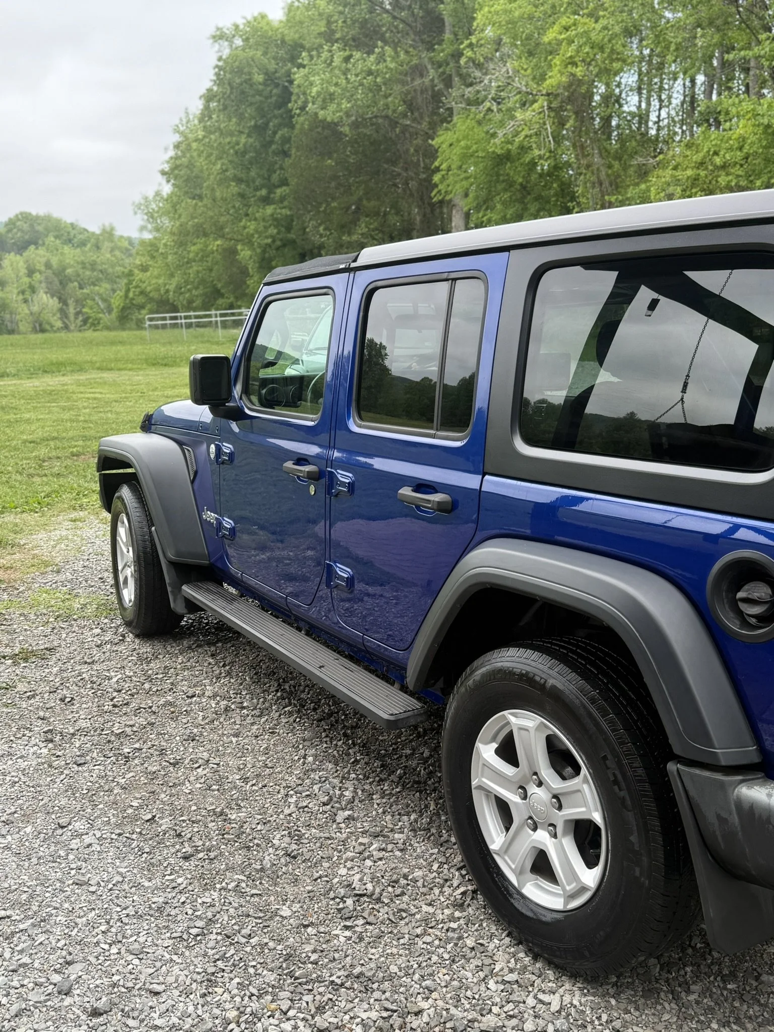 A blue Jeep SUV parked on a gravel area with a grassy field and green trees in the background.