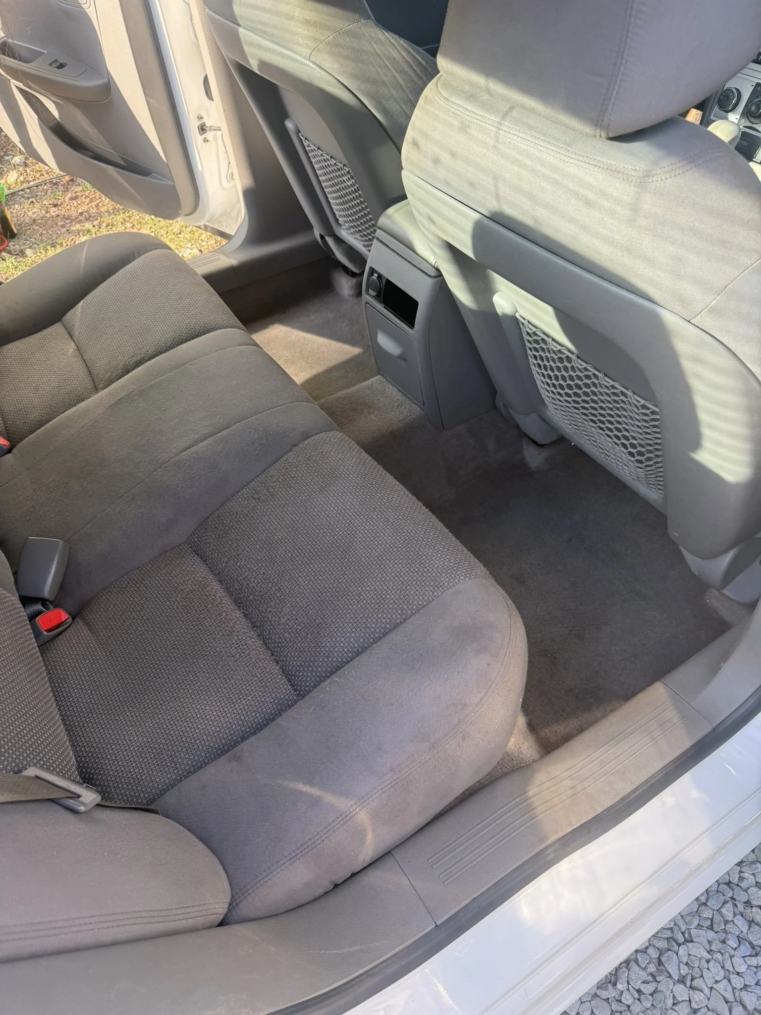 The interior of a car showing the back seat, front passenger seat, and part of the console. The seats are upholstered in gray fabric, and the car's door is open, showing the ground outside with gravel.