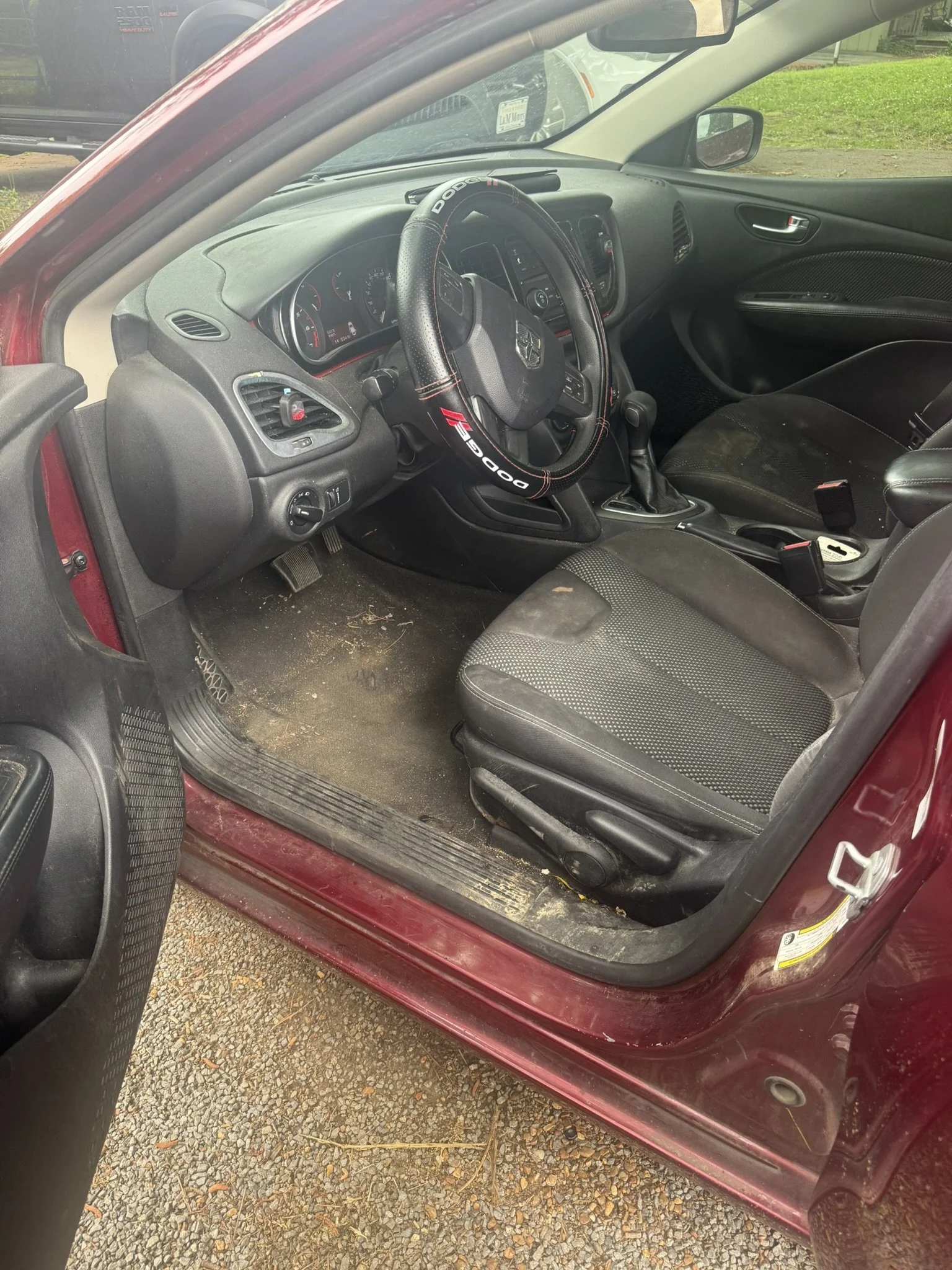 The interior of a red car showing the driver's side with a Dodge steering wheel, dashboard, gear shift, and seat, on a gravel surface with some grass outside.