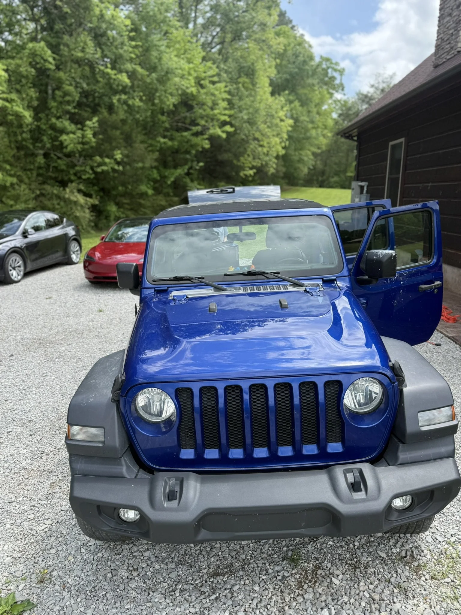 Front view of a blue Jeep Wrangler parked on a gravel driveway with other cars, a house, and trees in the background.