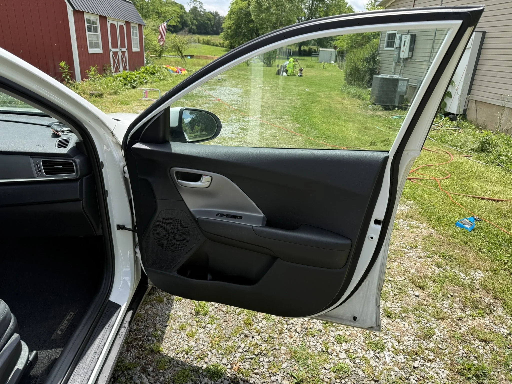 View from inside a car with an open driver-side door, showing a grassy backyard with a red barn, a small garden, and various outdoor items including a green golf bag and a blue equipment case.