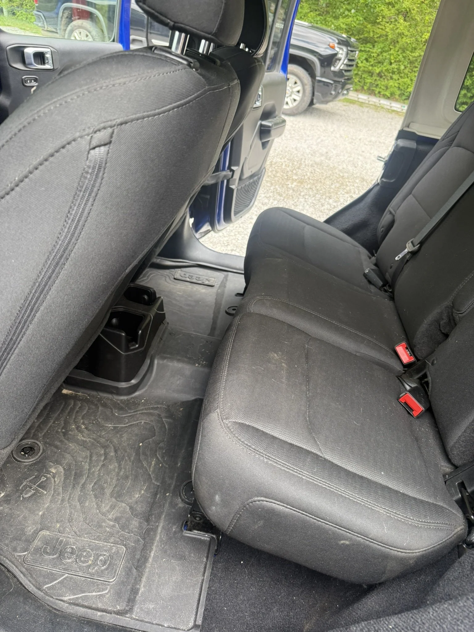 Interior of a Jeep vehicle showing the back seats, black fabric seats, and a rubber floor mat with the Jeep logo. Outside the vehicle, there is a parking lot with other cars and a green hedge.