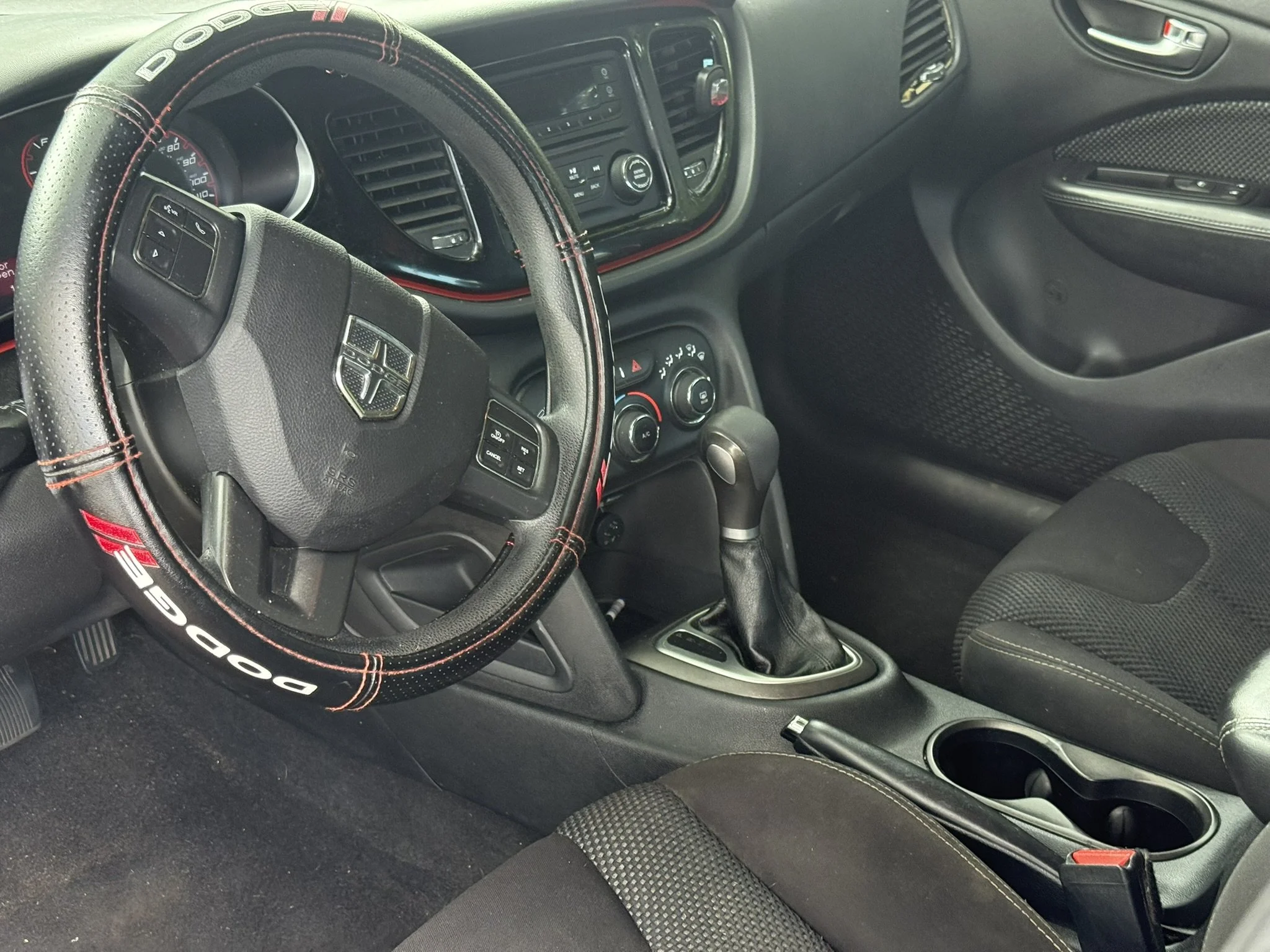 Inside view of a car dashboard showing a Dodge steering wheel with red stitching, speedometer, air conditioning controls, and a gear shift lever.