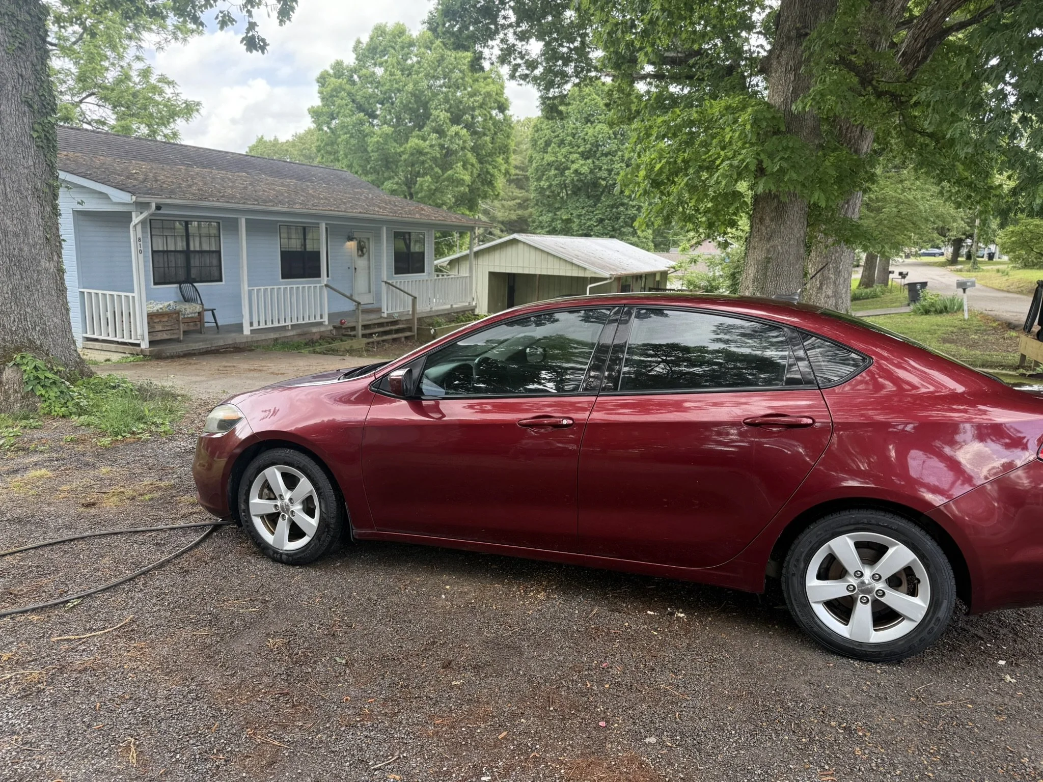 A red car parked on a gravel driveway in front of a light blue house with white trim and black window screens. Large green trees surrounding the house and driveway, with a smaller white building beside the house. The sky is partly cloudy.