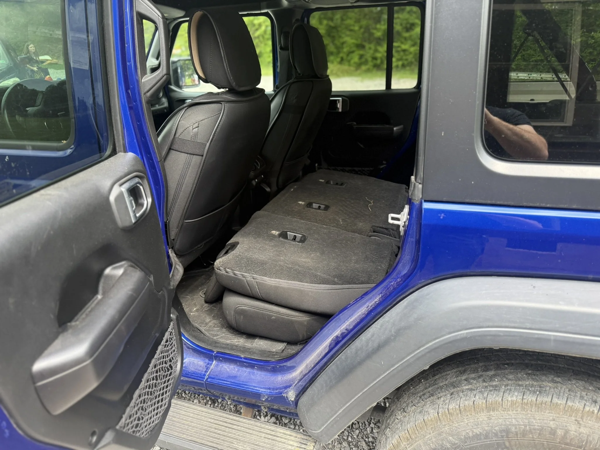 The interior of a blue SUV showing back seats folded down and front seats with headrests. The photo is taken through the open rear door, with a woman holding a tray visible in the reflection on the window.