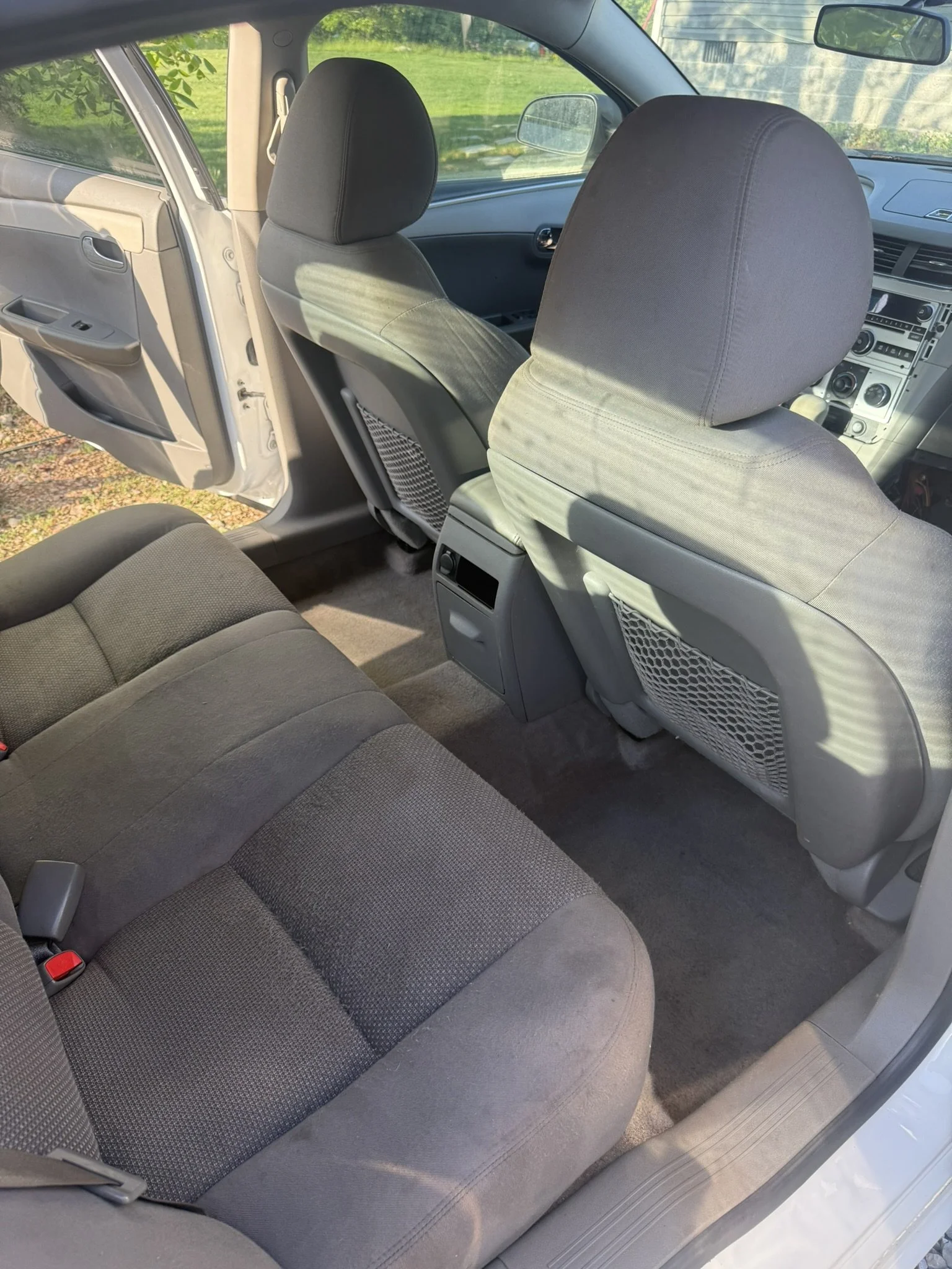 The backseat of a car with dark gray fabric seats, and the front seats are also visible with their headrests. Sunlight is shining into the vehicle, illuminating the interior. The door is open, revealing part of the outdoors with green grass and trees