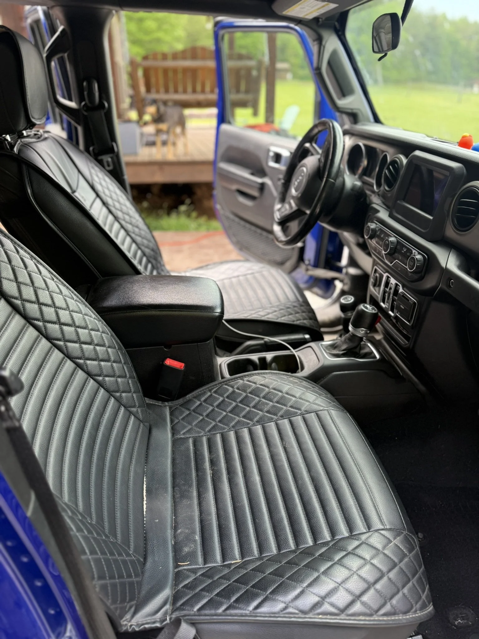 The interior of a vehicle showing the front seats with black and gray quilted seat covers, the dashboard, steering wheel, gear shift, and a view outside through the open door with a wooden porch and a dog in the background.