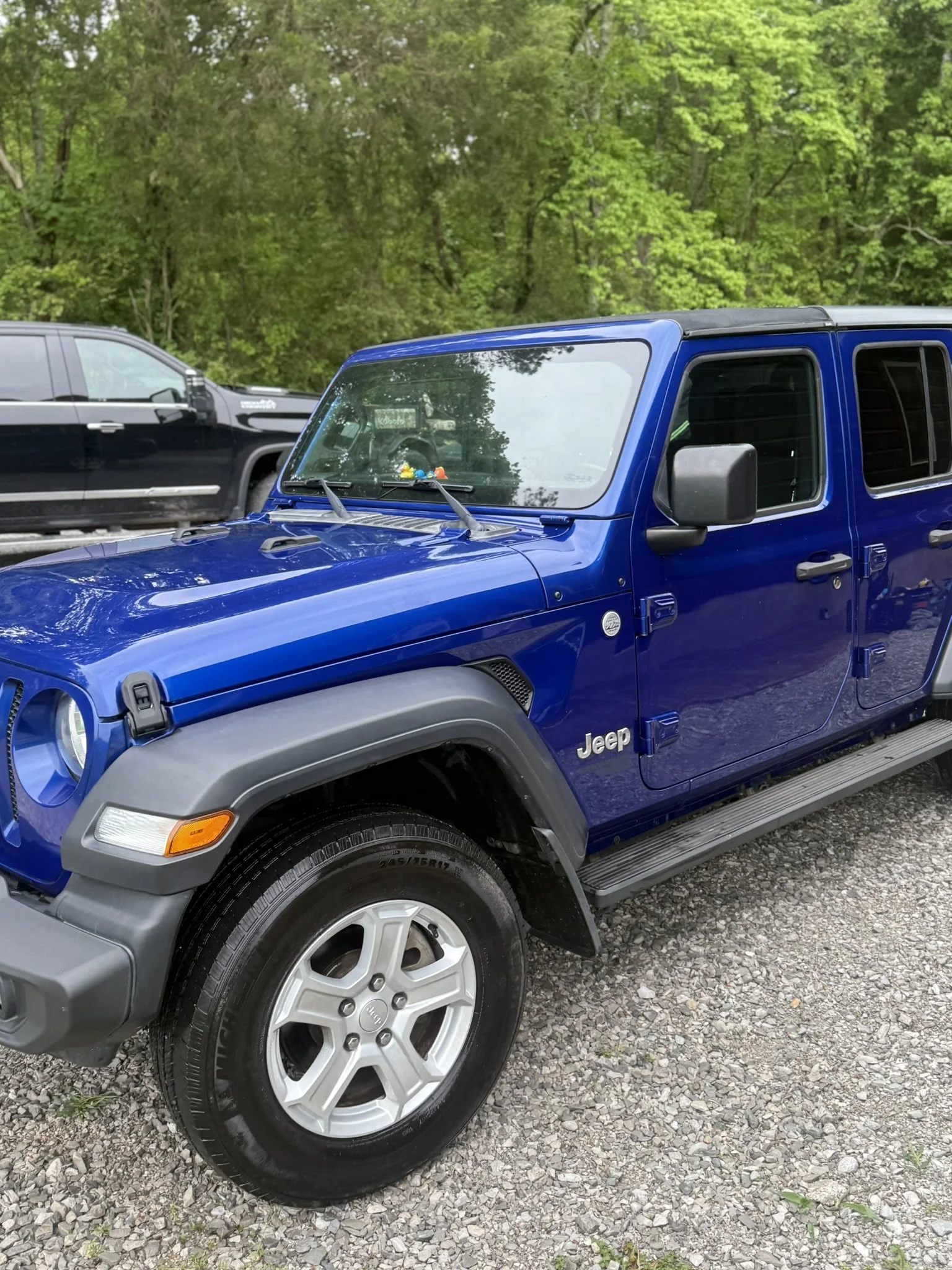 A blue Jeep vehicle parked on a gravel area with a second black vehicle parked behind it. In the background, there are green trees and foliage.