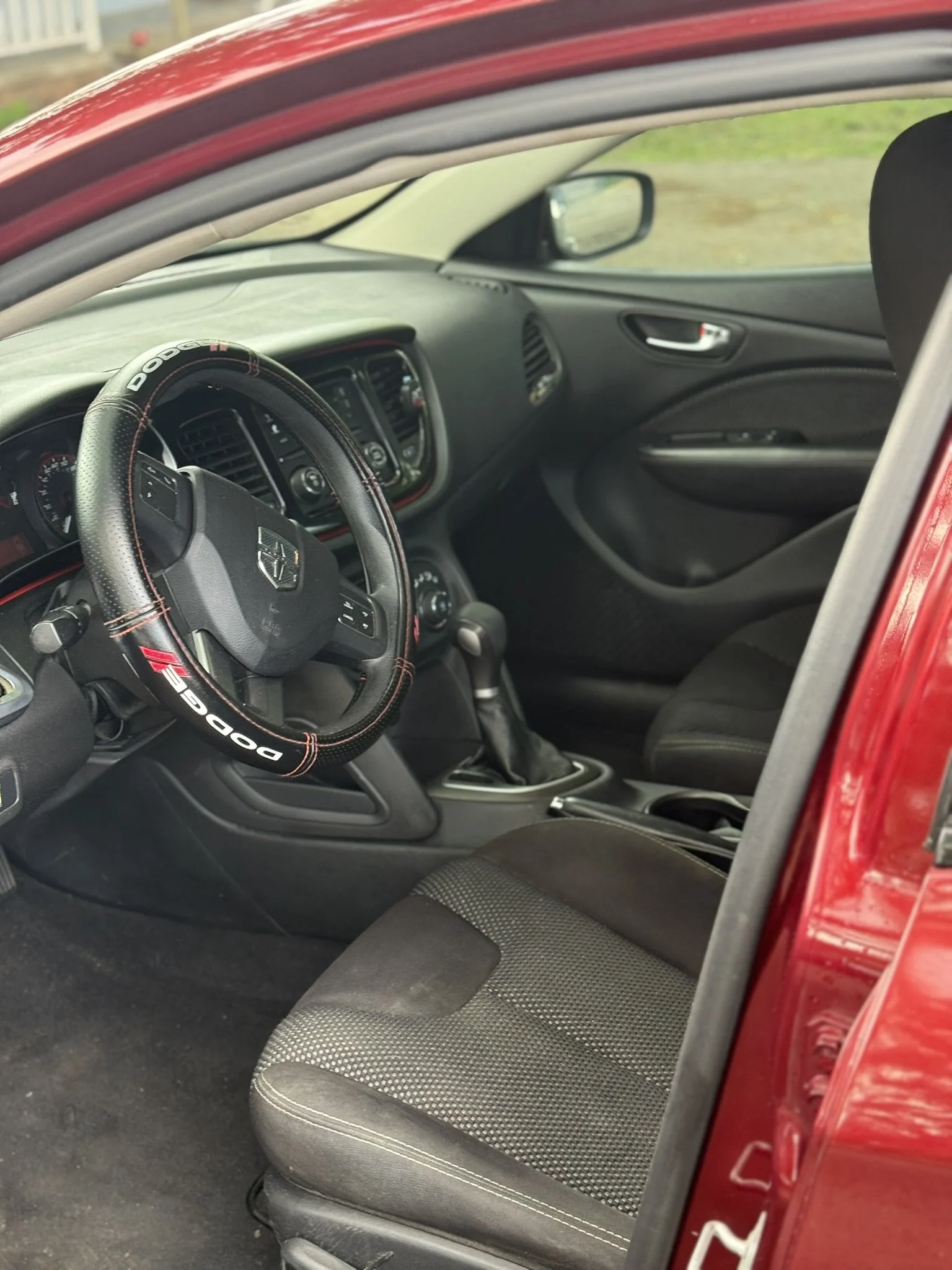 The interior of a red car showing the driver's seat, steering wheel with a Dodge logo, dashboard, and passenger seat.