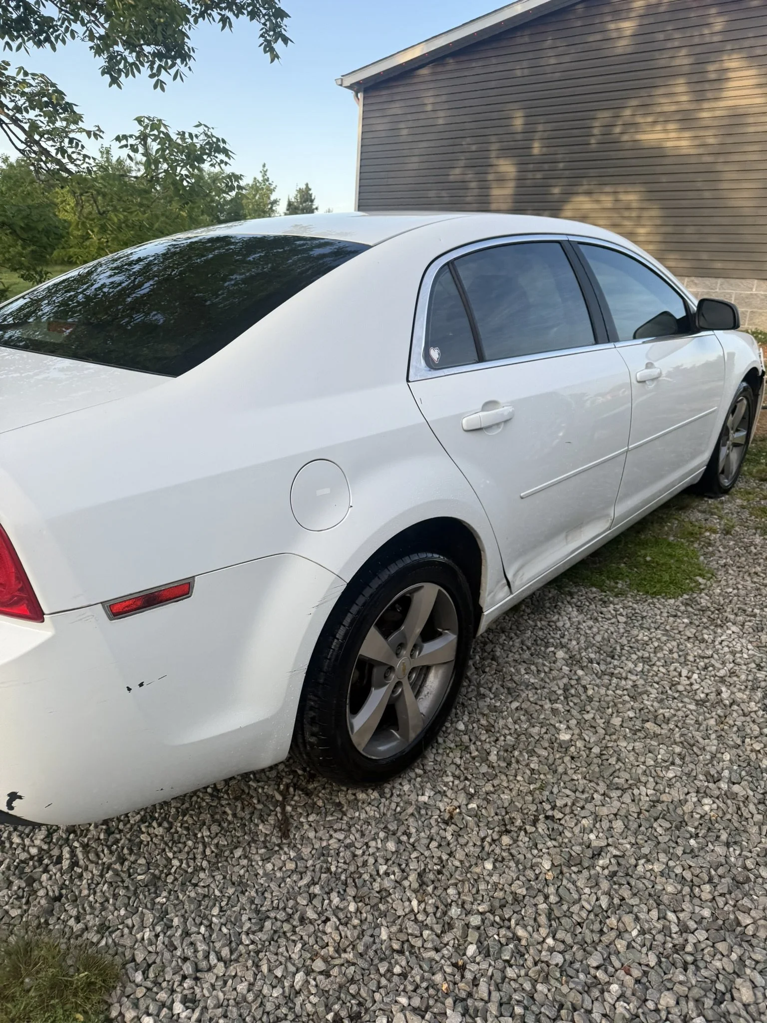 White sedan car parked on gravel with trees and a wooden building in the background.