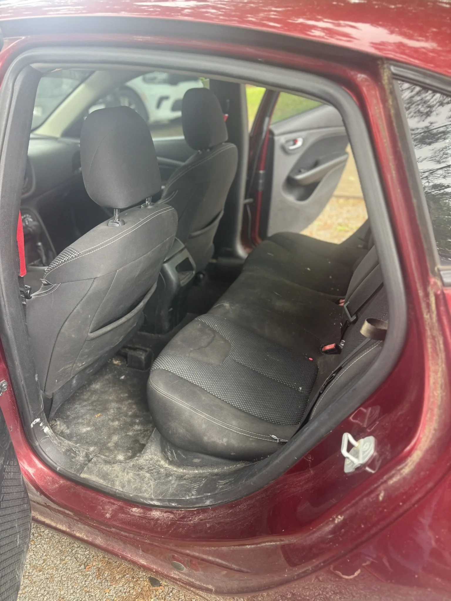 Inside view of a red car showing the backseat, with black fabric seats, and the front seats with headrests. The car door is open, revealing the interior and part of the outside ground.