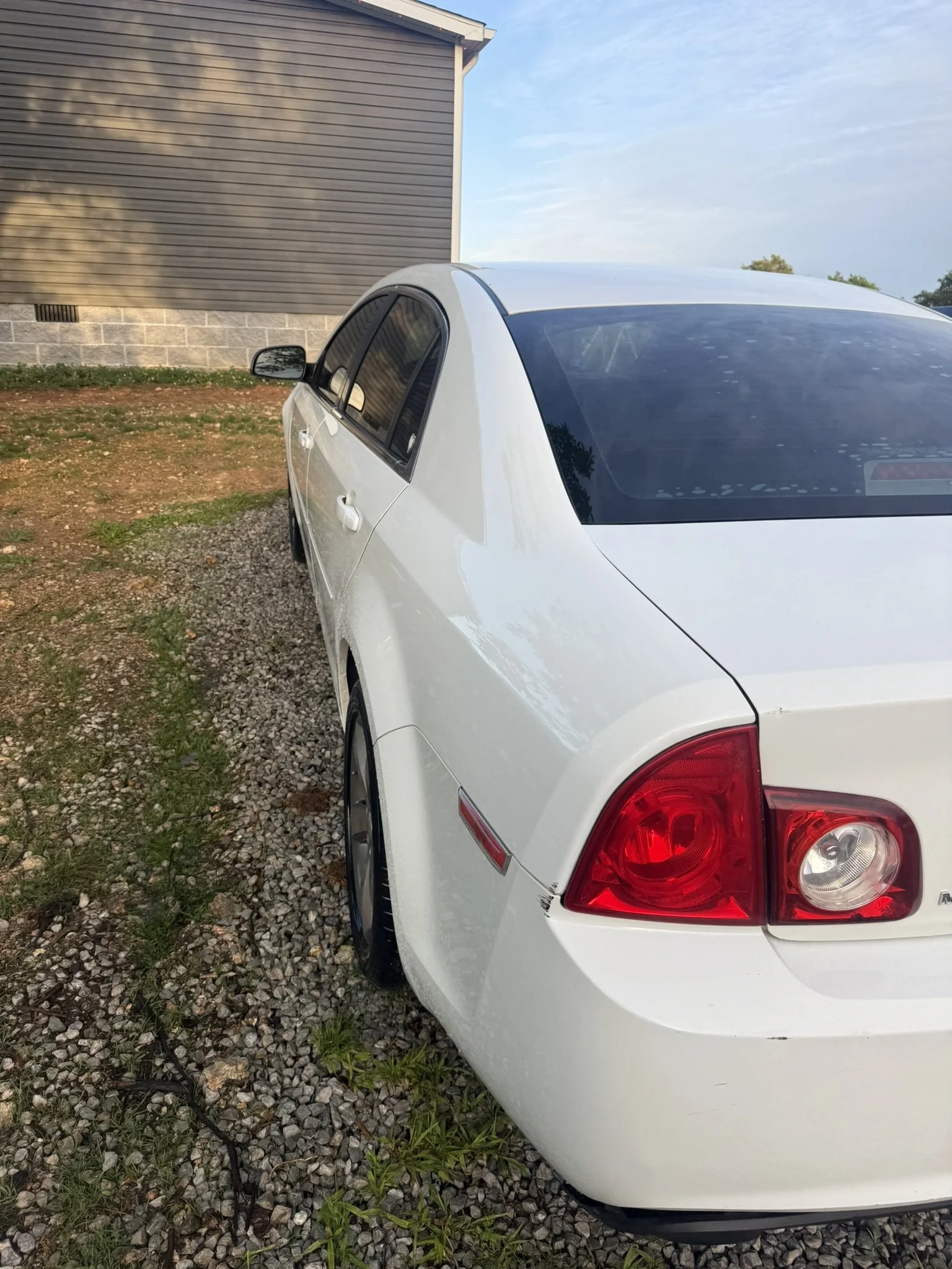 A white sedan car parked on gravel ground next to a house with brown siding and a brick foundation, under a blue sky.