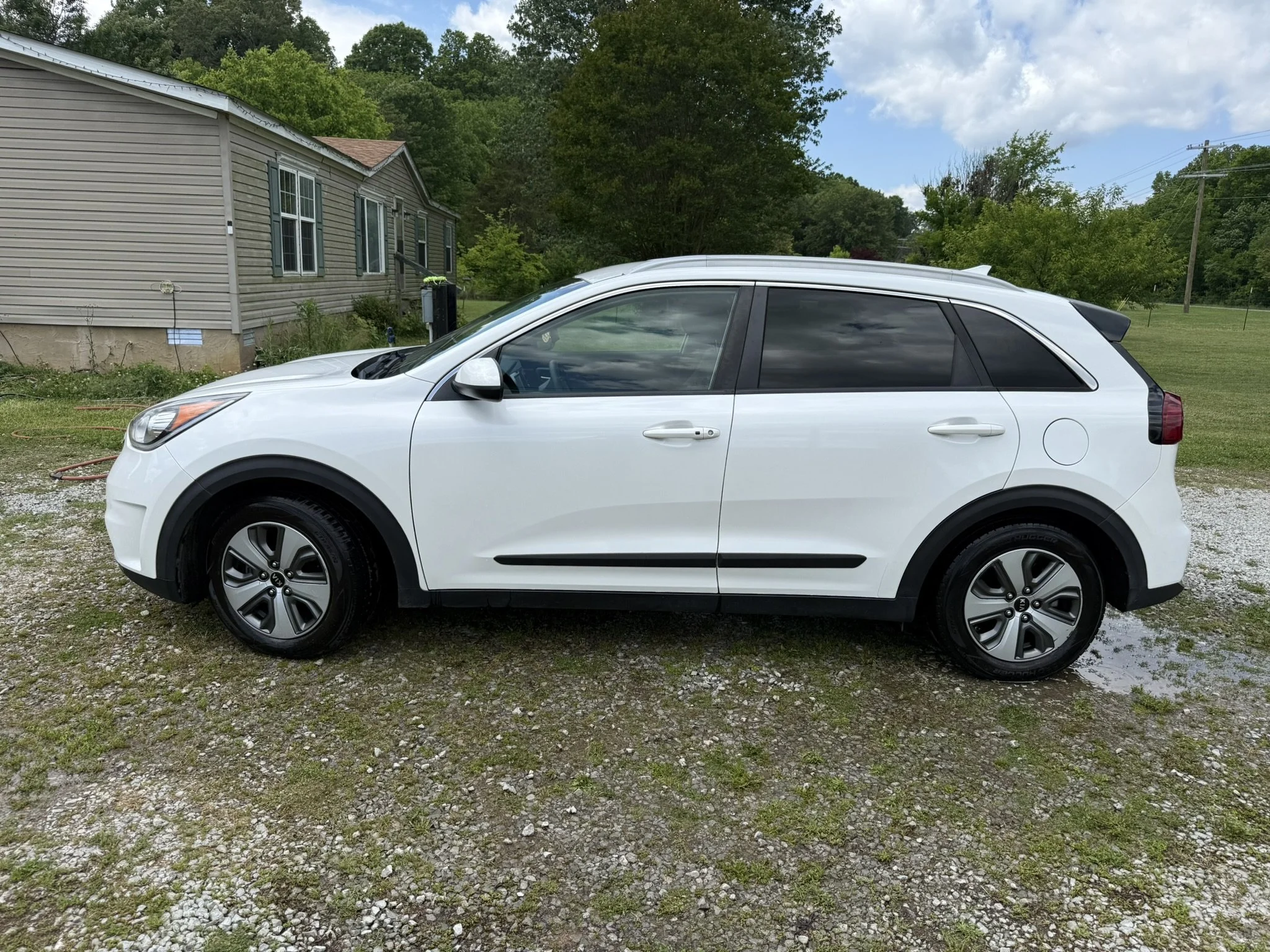 A white SUV vehicle parked on a gravel driveway in front of a house with beige siding, green shutters, and a slightly sloped roof surrounded by trees and a lawn, under a partly cloudy sky.