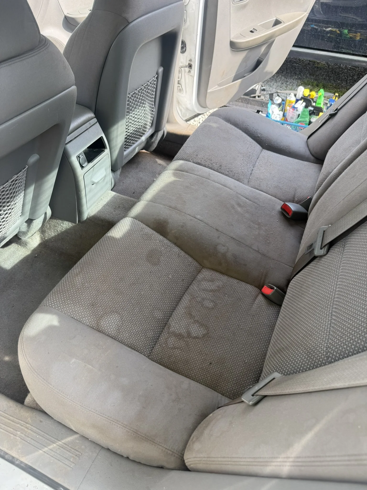 Backseat of a car showing gray fabric seats, seat belts, and rear of front seats with mesh pockets, with a view through the open rear door to cleaning supplies outside.